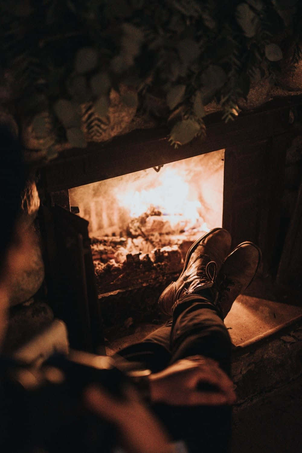 Cozy Fire 桌布 壁紙 — 舒適的雙腳穿著靴子，在光線昏暗的小屋中，在綠植環繞的噼啪作響的壁爐前休息。