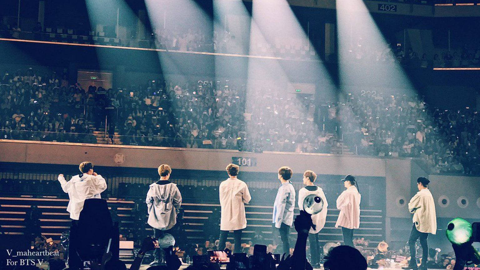 舞台上的BTS可愛風 壁紙 — 七位表演者站在燈光璀璨的舞台上，面對擠滿場館的龐大觀眾，在戲劇性的聚光燈下演出。