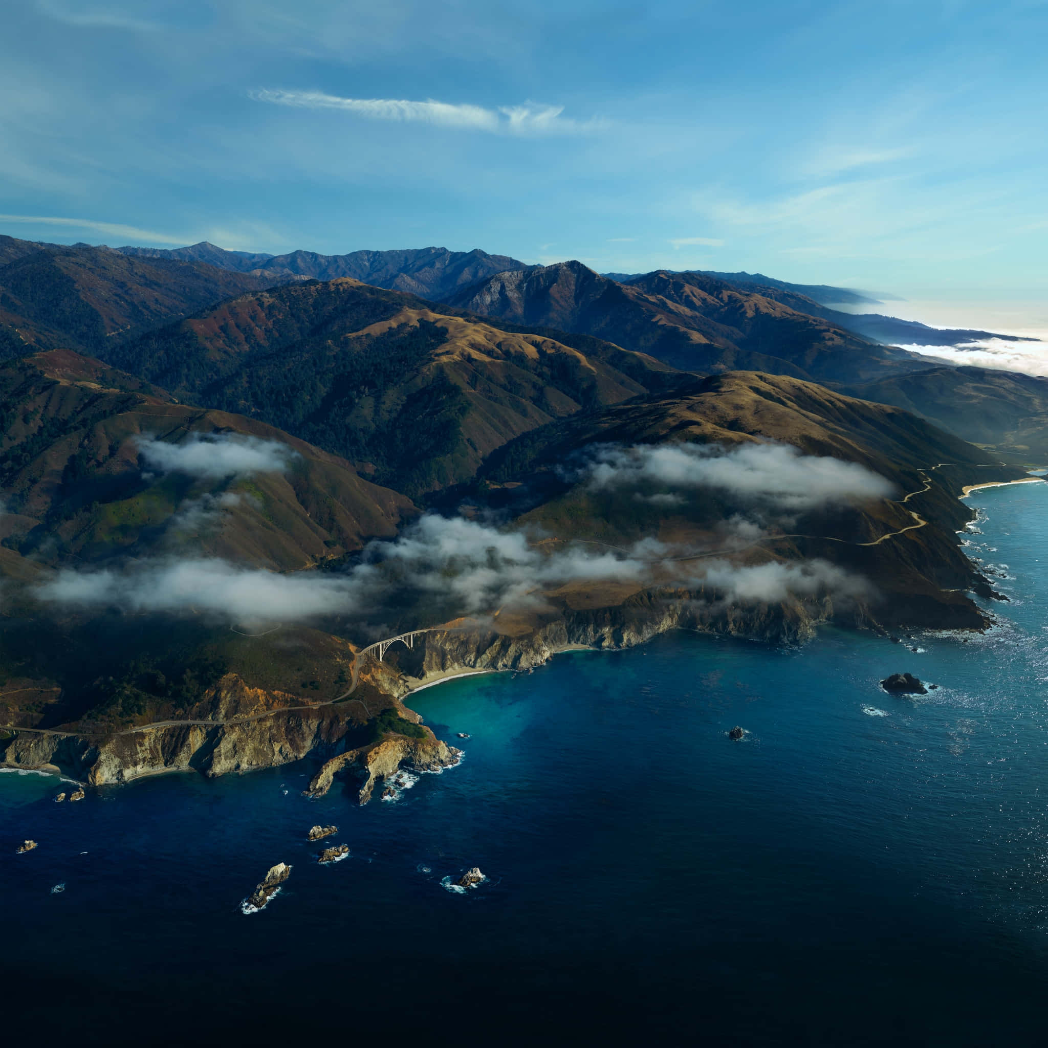 Big Sur 桌布 壁紙 — 航拍視角，一條蜿蜒的沿海高速公路緊貼崎嶇懸崖，伴隨碧綠海洋與起伏山丘，在清澈藍天下延伸。