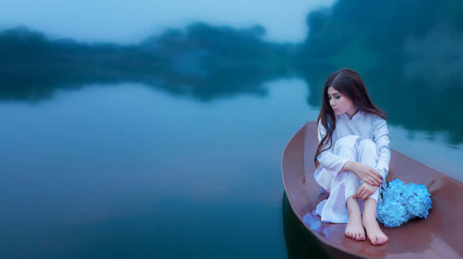 船上的青春少女 壁紙 — 一位身穿白衣的寧靜女子赤腳坐在小船中，在黃昏平靜霧氣瀰漫的水面上手持藍色繡球花。