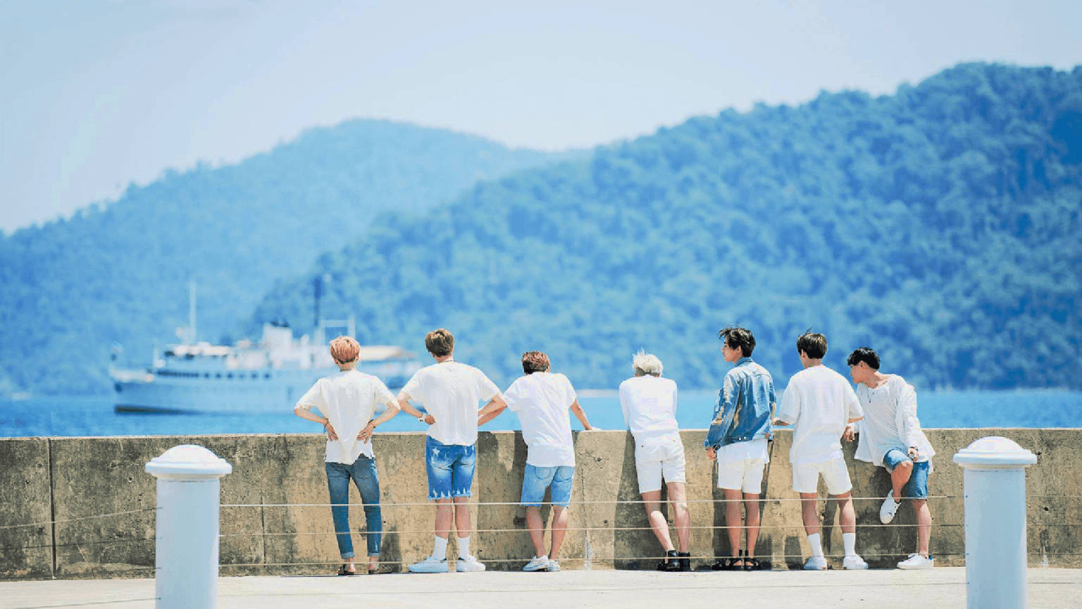 船邊的BTS可愛風 壁紙 — 七名身穿白襯衫和短褲的男子站在碼頭欄杆上，凝視著一艘遊輪在明亮的藍天下駛過鬱鬱蔥蔥的綠色山丘。