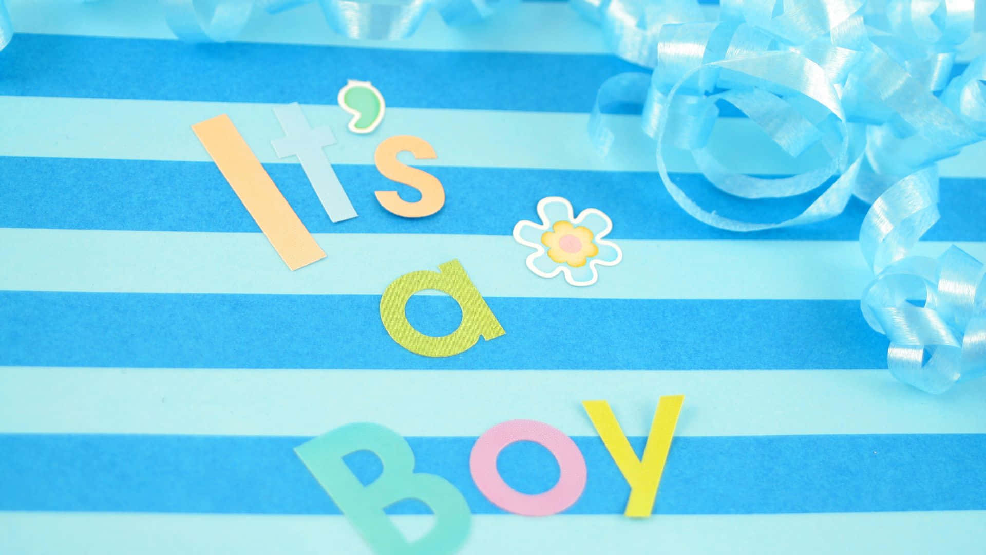 Boy 桌布 壁紙 — 色彩繽紛嘅紙字母拼出 'It's a Boy'，放喺藍色間條紙上面，旁邊有藍色絲帶同花貼紙。