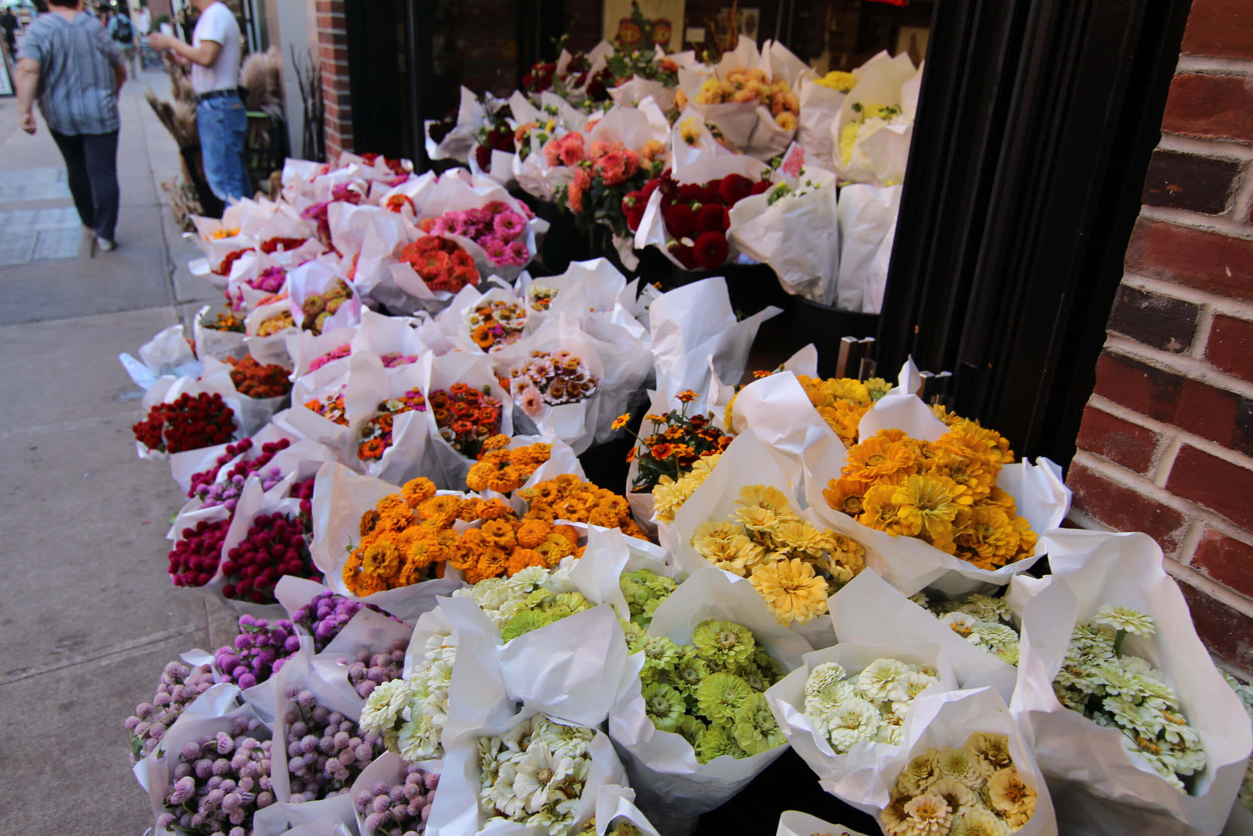 Flower Market 桌布 壁紙 — 色彩繽紛嘅花束用白紙包住，排列喺繁忙城市行人路嘅磚牆店舖前面。