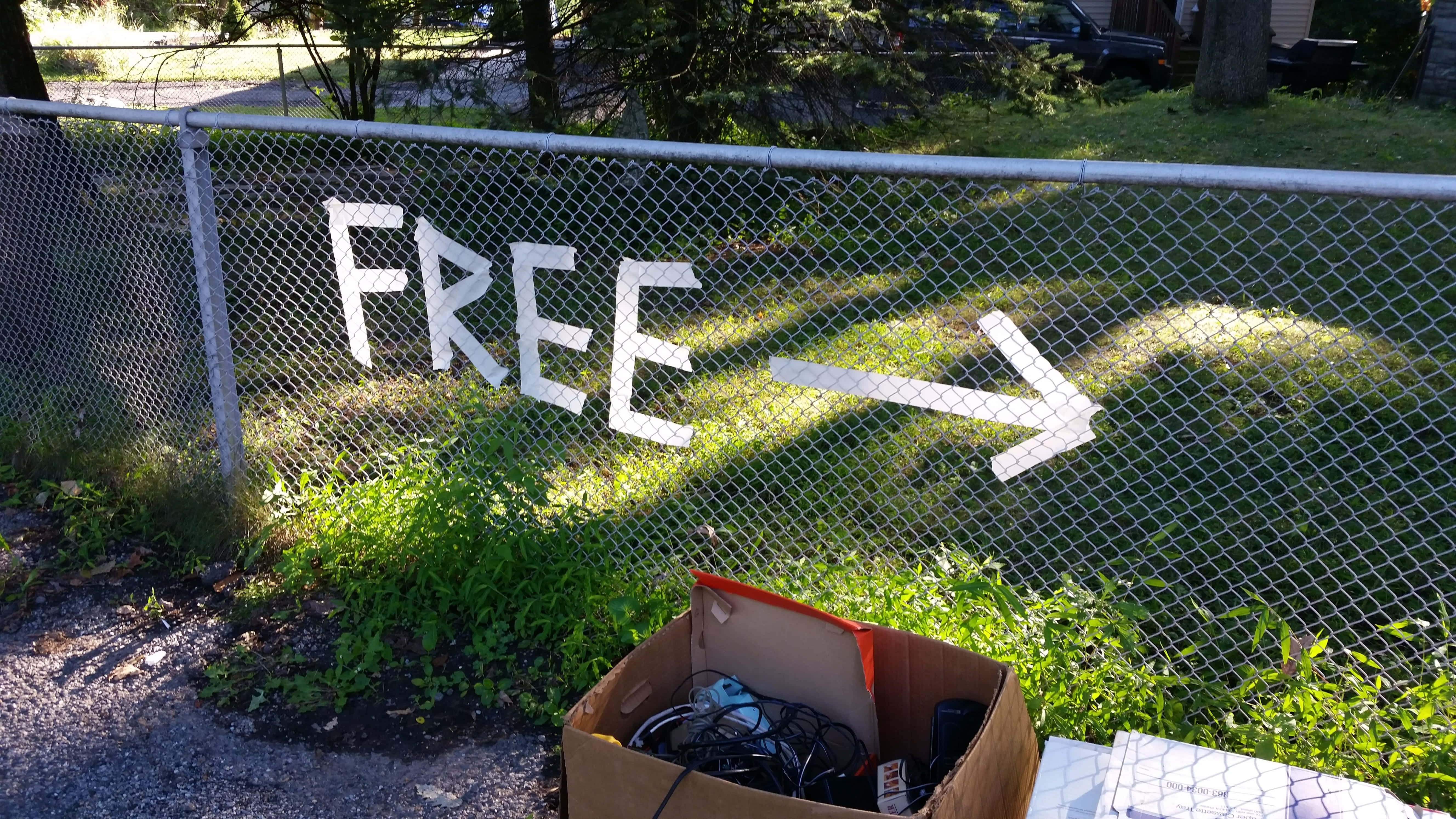色彩繽紛的二手市集場景 壁紙 — 一個鐵絲網圍欄上畫有「FREE」和一個箭頭，指向一個放在草地上的紙箱，裡面裝滿了廢棄物品。
