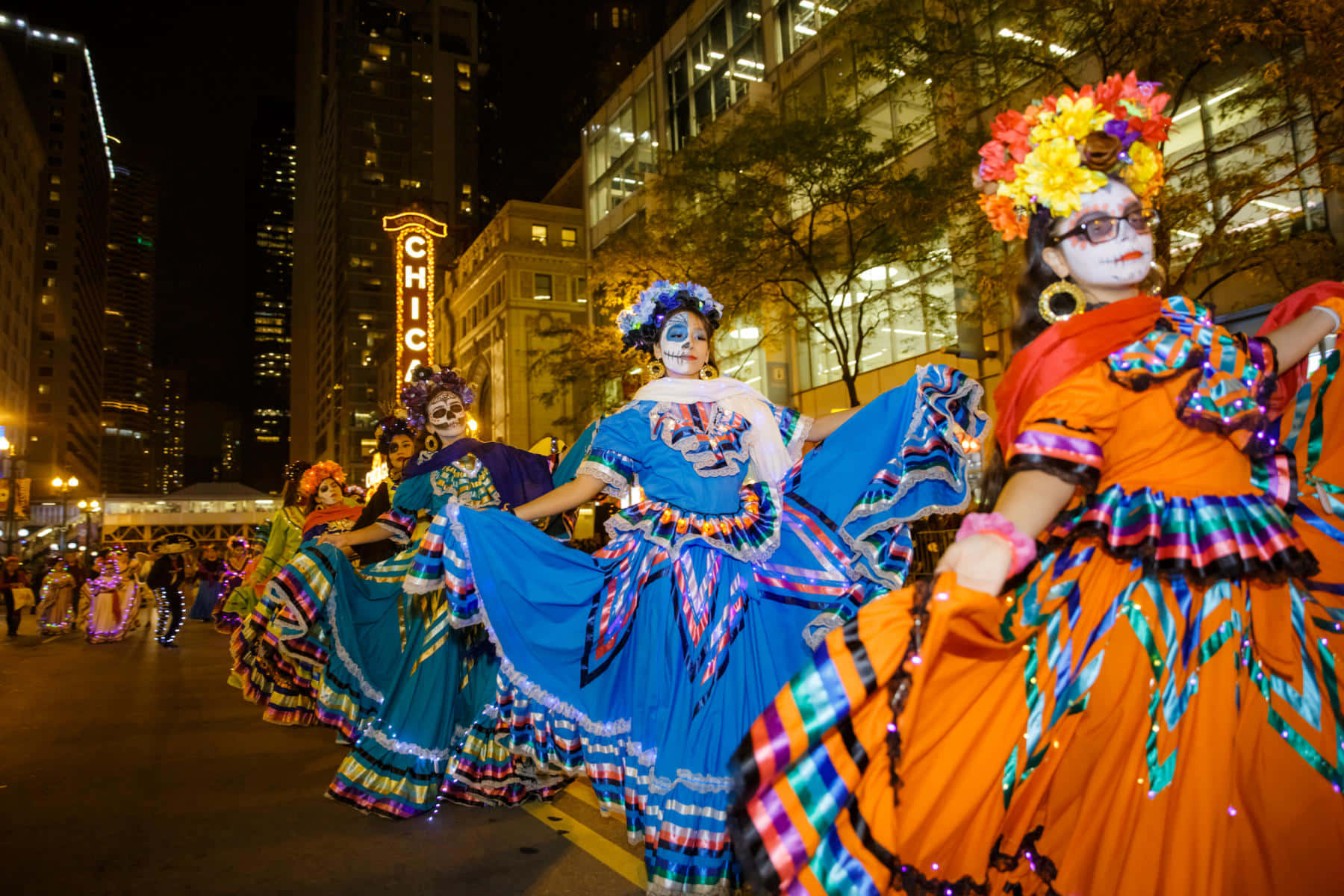 Halloween Parades 桌布 壁紙 — 色彩繽紛的傳統服飾舞者在夜晚城市燈光下表演，背景中 'CHICAGO' 標誌閃爍發光。