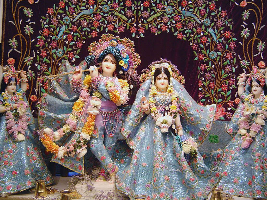 krishna god 桌布 壁紙 — 色彩繽紛的印度教神祇克里希納和拉達，身著華麗藍色服飾，周圍環繞著花卉裝飾和侍從。