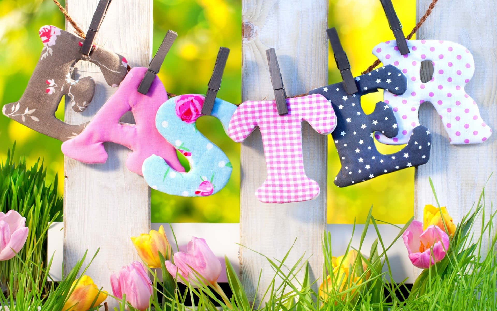 Happy Easter 桌布 壁紙 — 色彩繽紛的布料字母拼出 'EASTER'，掛在木柵欄上，下方是盛開的粉紅色和黃色鬱金香，在充滿活力的春季草地上。