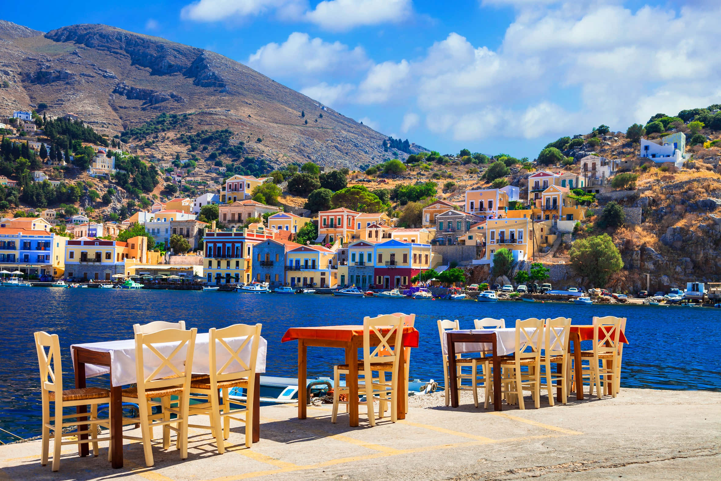 Greek Beach 桌布 壁紙 — 色彩繽紛的懸崖村莊房屋俯瞰著平靜的藍色大海，前景有戶外用餐桌，在明亮的陽光下。
