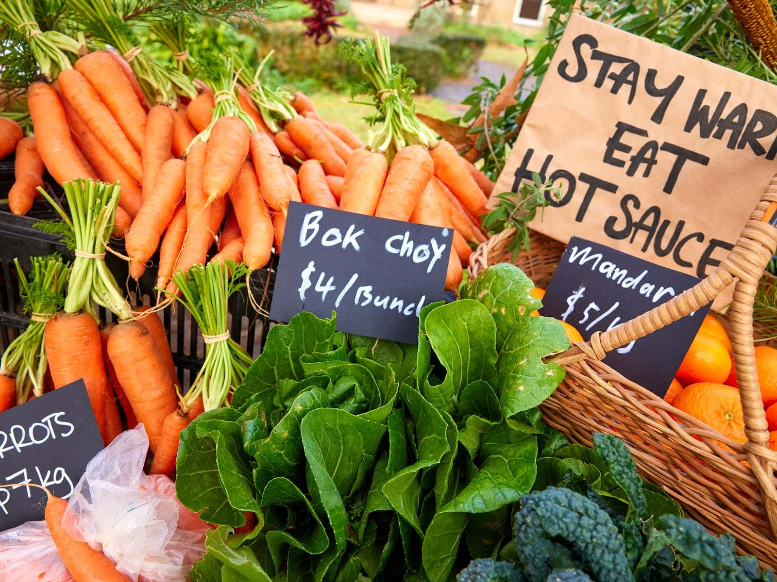 Farmers Market 桌布 壁紙 — 色彩繽紛的新鮮農產品，包括紅蘿蔔、白菜和橙子，在戶外農貿市場展示，附有手寫價格標籤。