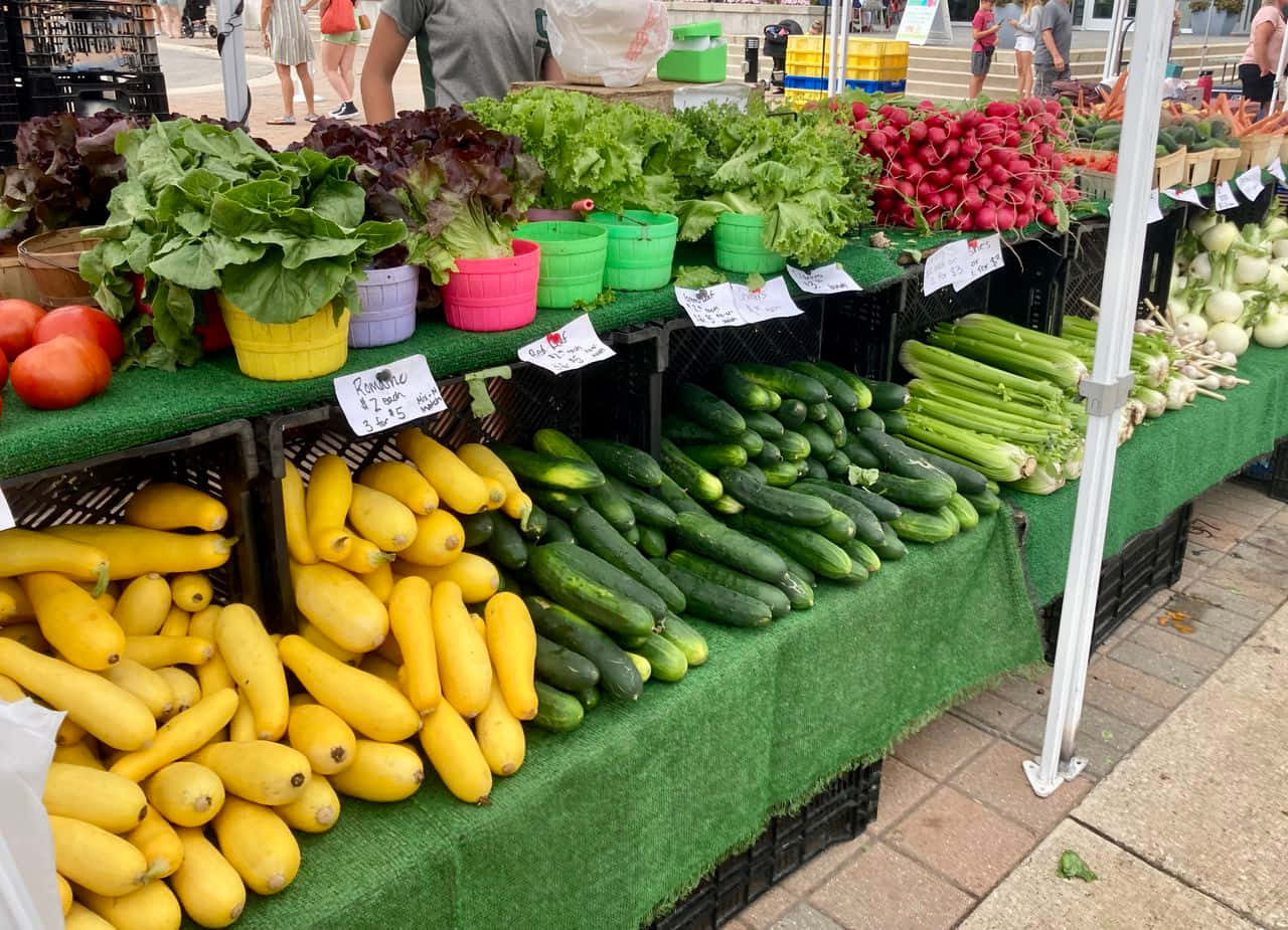 Farmers Market 桌布 壁紙 — 色彩繽紛的新鮮農產品，如黃色南瓜、青瓜和蘿蔔，在戶外農夫市場攤位的彩色籃子中展示。
