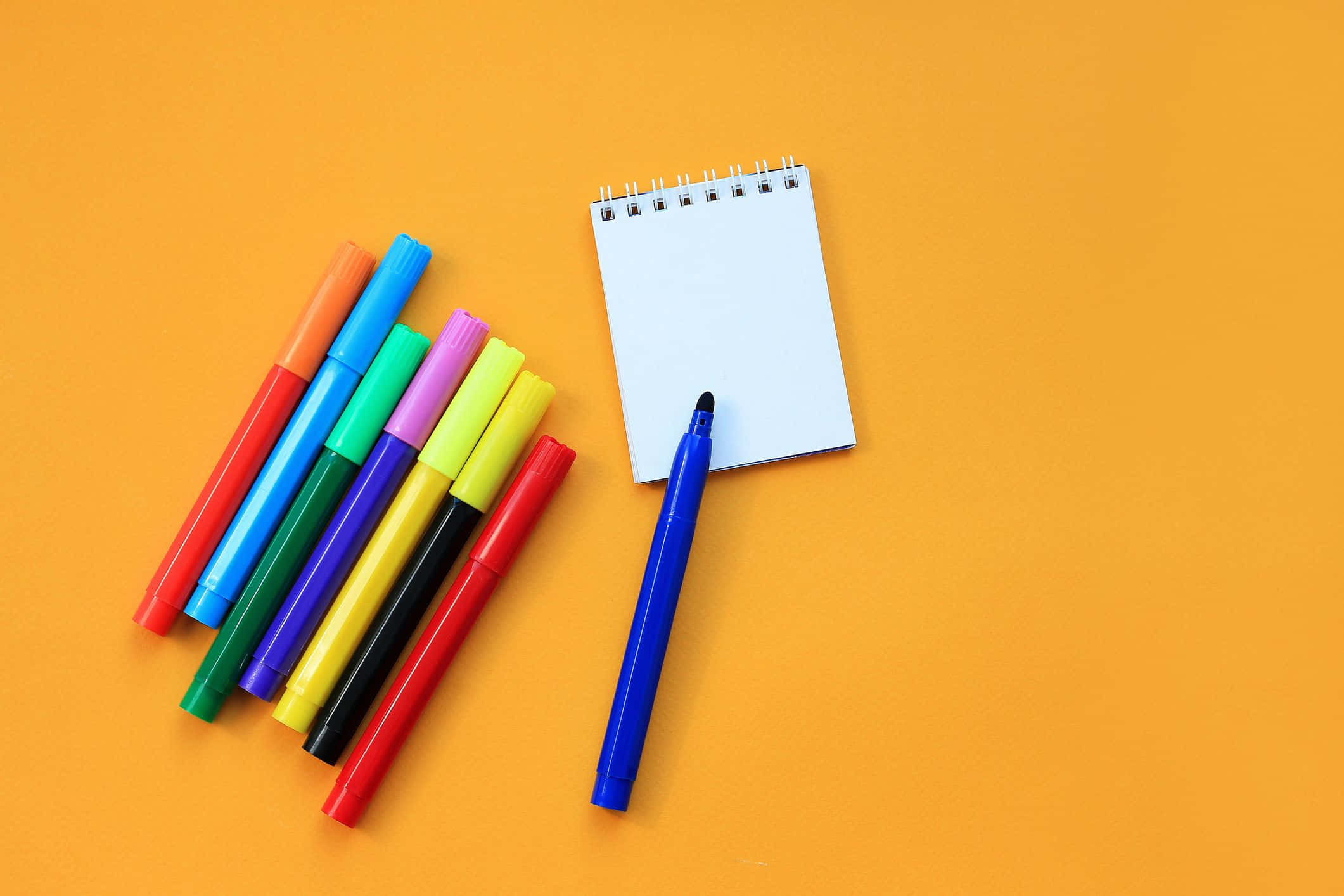 pencil 桌布 壁紙 — 色彩繽紛的標記筆和空白螺旋筆記本在溫暖的橙色背景上排列。