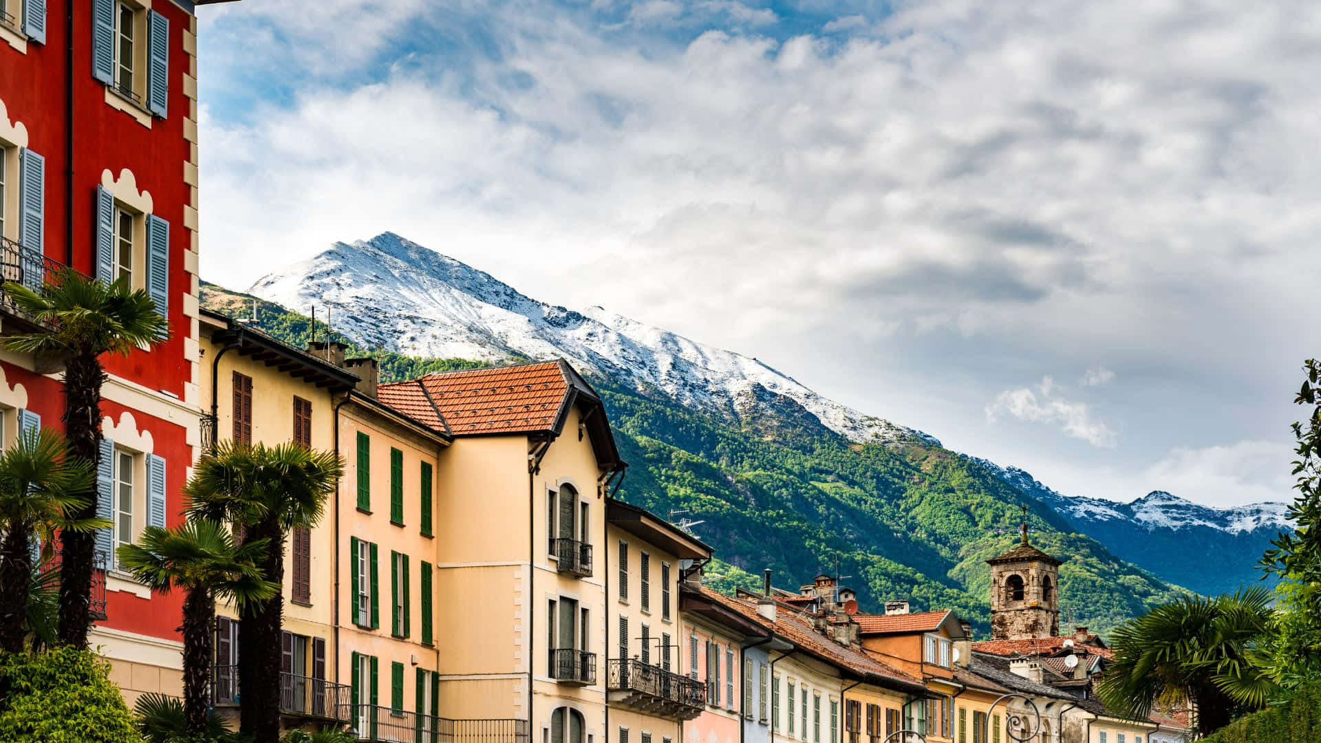 Switzerland 桌布 壁紙 — 色彩繽紛的歐洲聯排別墅排列在街道上，背後是白雪覆蓋的山脈，天空部分多雲。