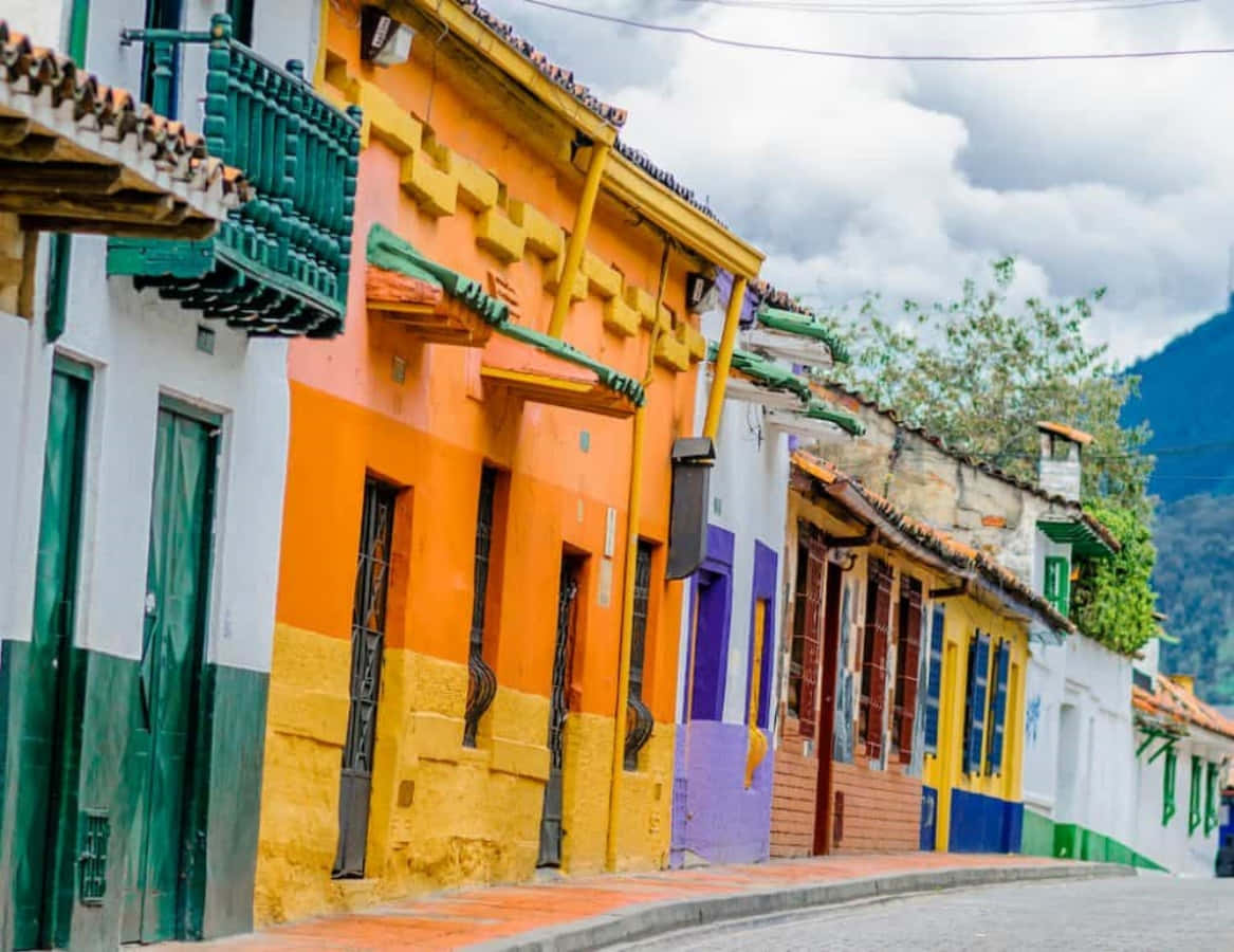 Colombia 桌布 壁紙 — 色彩繽紛的殖民時期房屋排列在一條寧靜的鵝卵石街道上，背景是山脈，天空多雲。