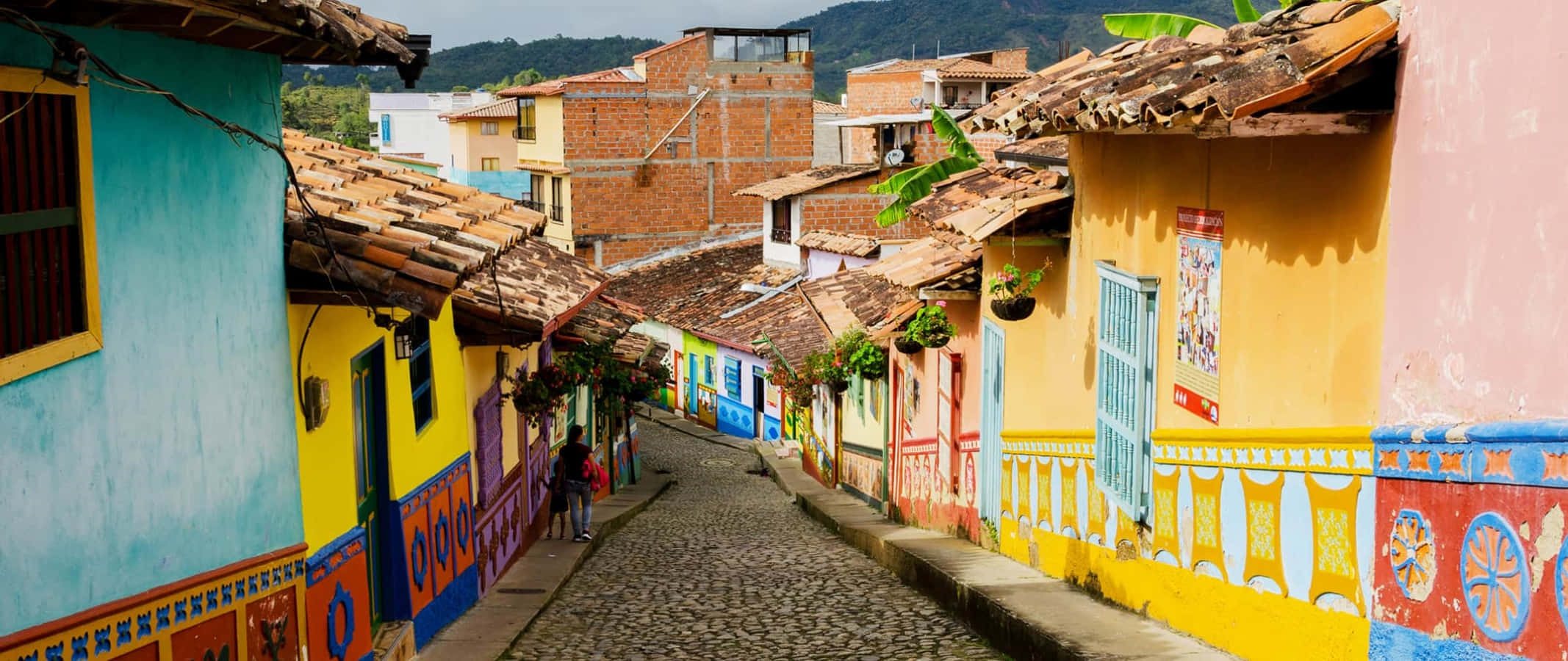 Colombia 桌布 壁紙 — 色彩繽紛的殖民風格房屋排列在明亮的日光下山腰村莊的一條狹窄鵝卵石街道上。