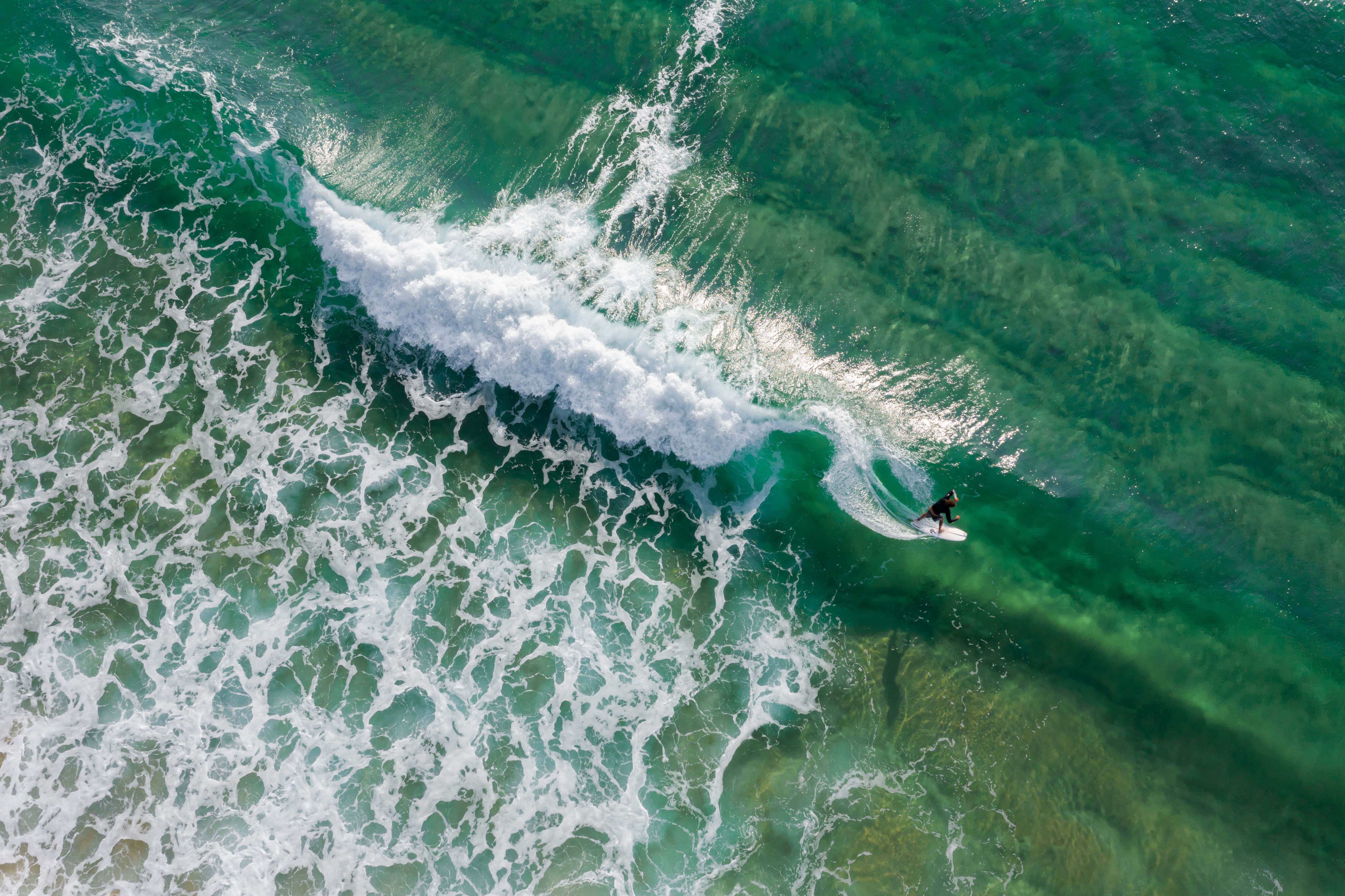 色彩繽紛的氣球在戶外飄浮 壁紙 — 從空中俯瞰，一名衝浪者騎乘在碧綠色的海浪上，白色泡沫與清澈的綠色深處形成對比。