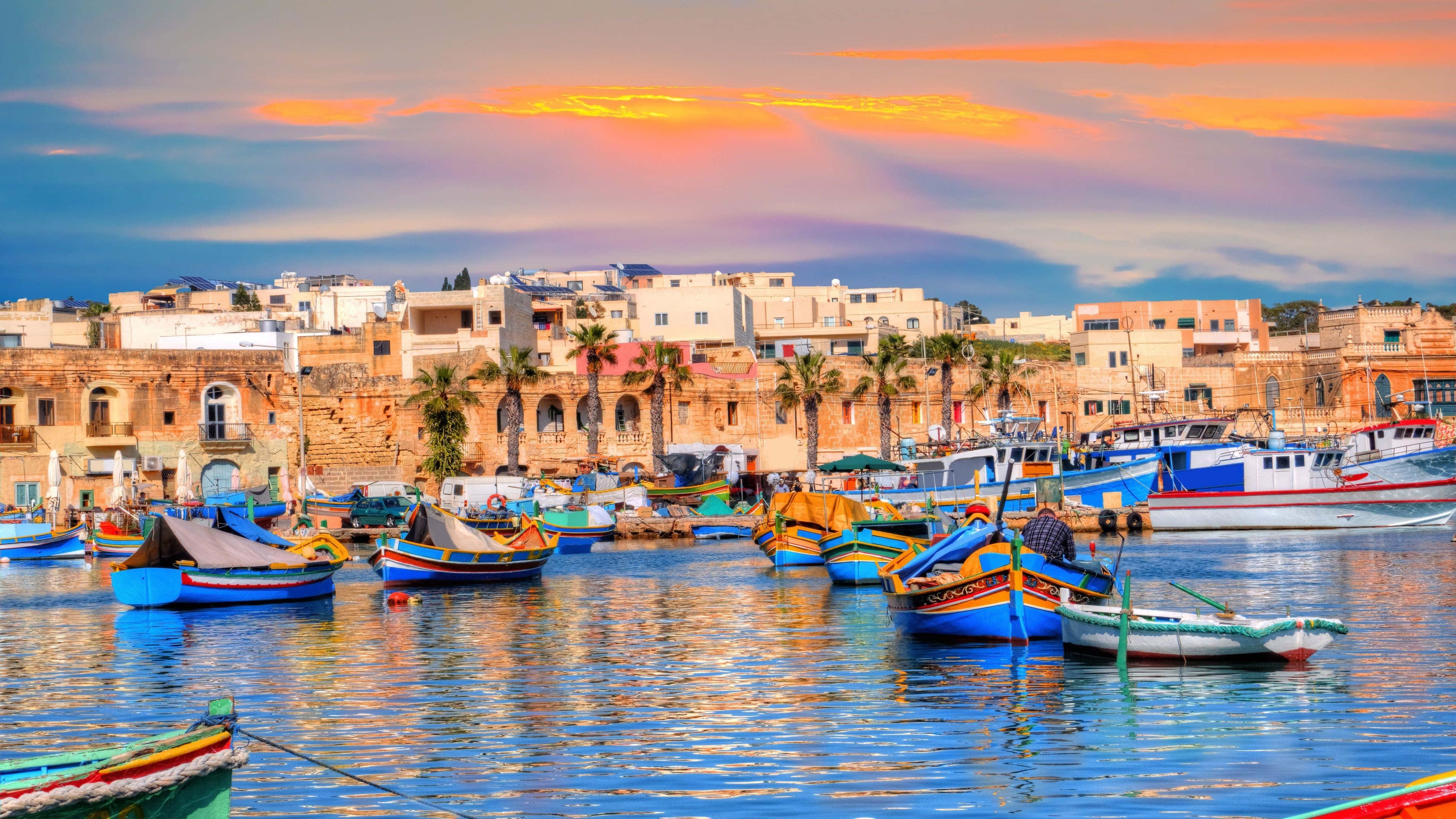 Malta 桌布 壁紙 — 色彩繽紛的漁船在日落時分漂浮在港口，背景有歷史悠久的石建築和棕櫚樹。