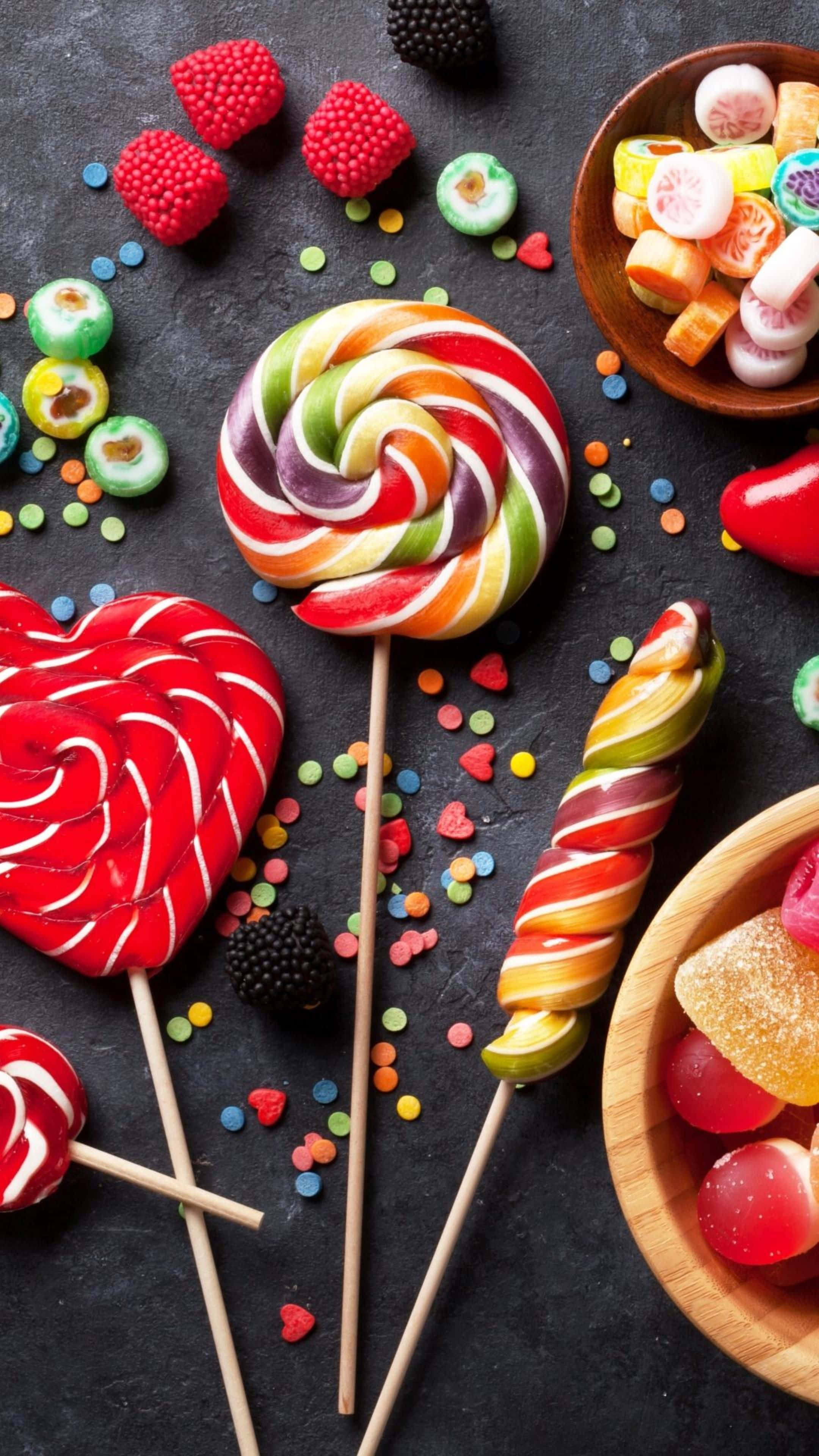 Food Photography 桌布 壁紙 — 色彩繽紛的漩渦棒棒糖和各式糖果散落在深色石板表面上，綴以彩色糖粒和新鮮漿果。