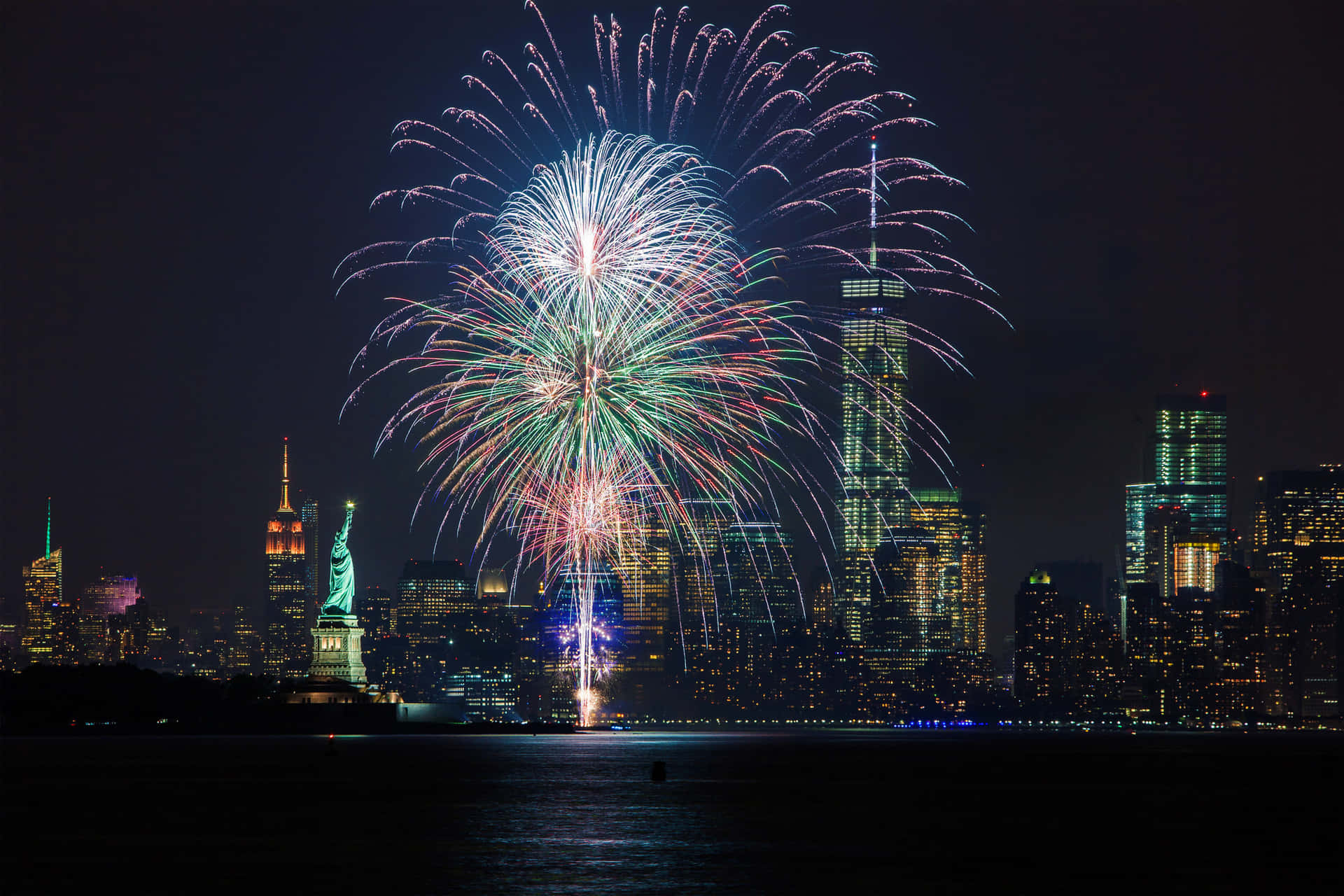 新年動畫 桌布 壁紙 — 色彩繽紛的煙火在夜間紐約市天際線上空綻放，自由女神像和燈火通明的摩天大樓倒映在水中。