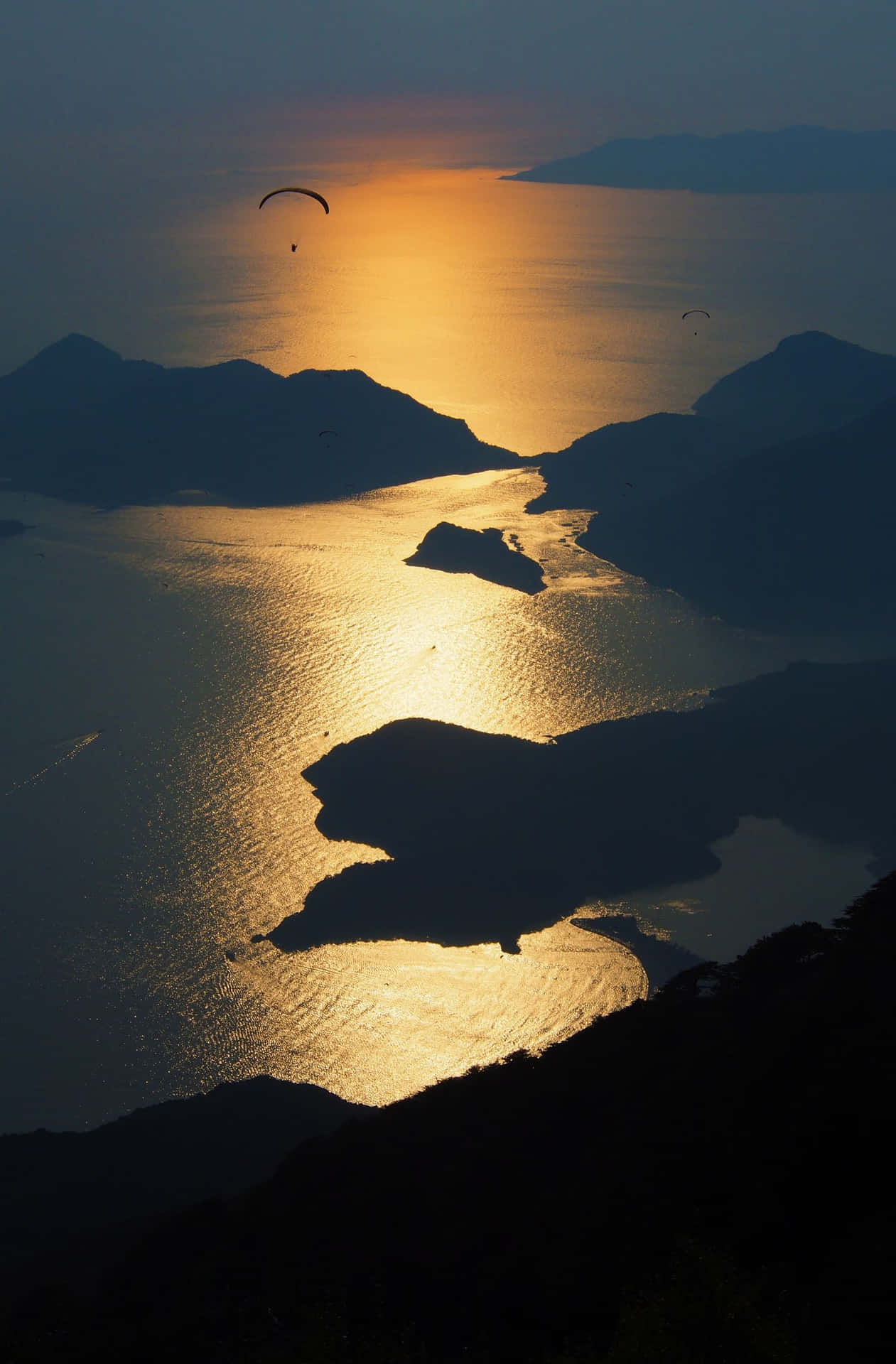 滑翔傘在夕陽下飛越金色海灣，伴隨著剪影般的島嶼和山丘。