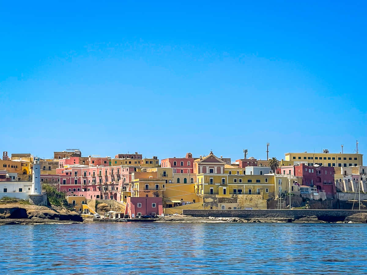 Italian Island 桌布 壁紙 — 色彩繽紛的粉彩建築依附在岩石海岸線上，下方是平靜的水面，上方是清澈的藍天。