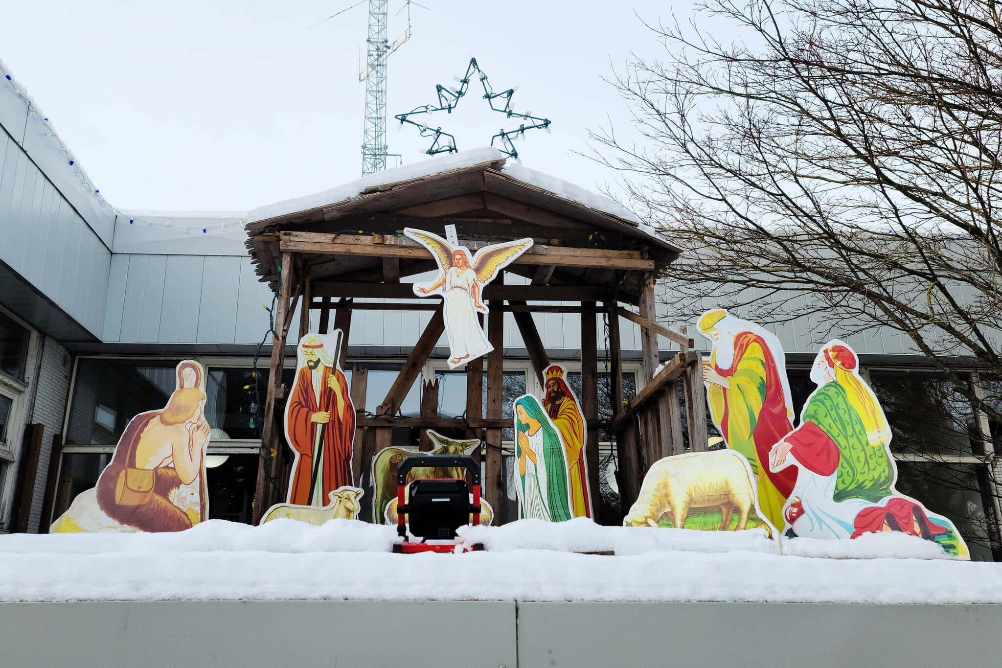 Nativity Scene 桌布 壁紙 — 色彩繽紛的紙板耶穌誕生人偶站在雪地上，上方有一個木製馬廄，頂部有星星，背景是光禿禿的樹木。