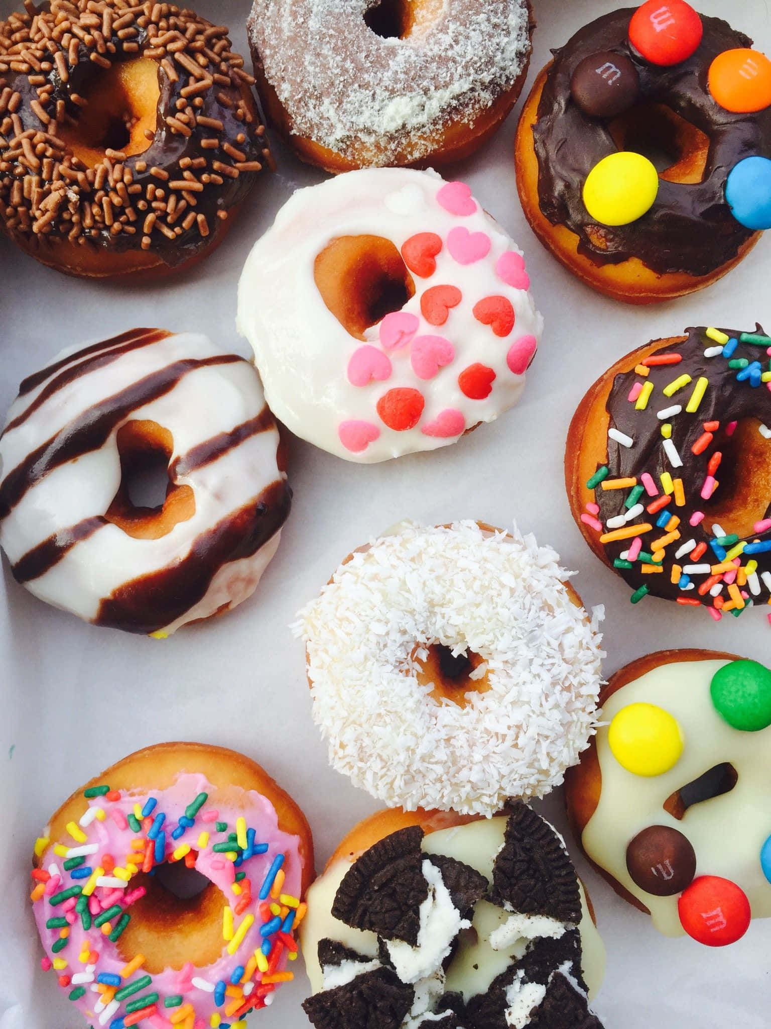 donuts 桌布 壁紙 — 色彩繽紛的釉面及裝飾甜甜圈，配上糖針、糖果和朱古力配料，整齊排列在白色牛油紙上。