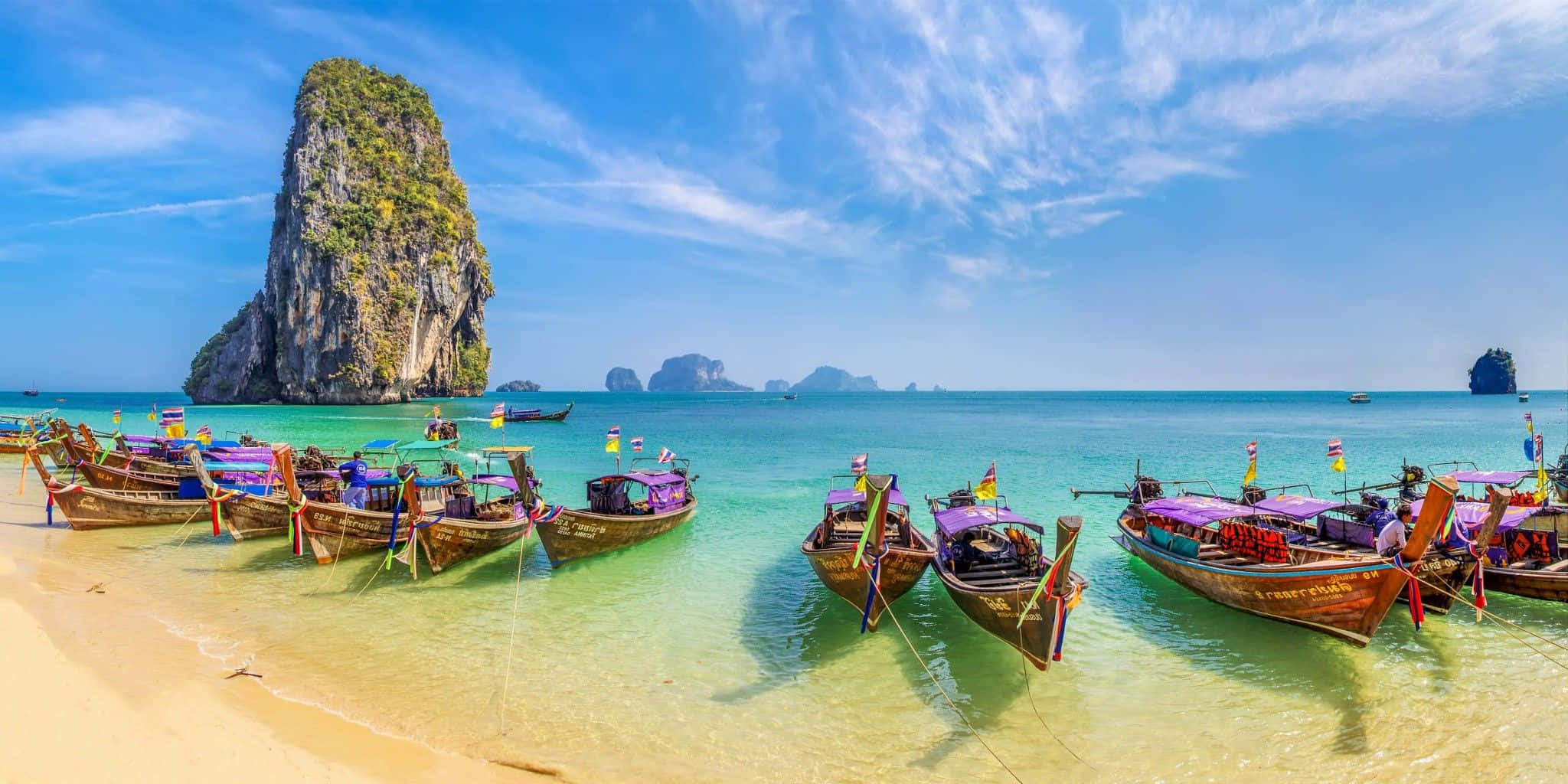 Boating 桌布 壁紙 — 色彩繽紛的長尾船停泊在金色沙灘旁，碧綠海水映襯下，高聳的石灰岩懸崖在明亮的藍天下巍然聳立。
