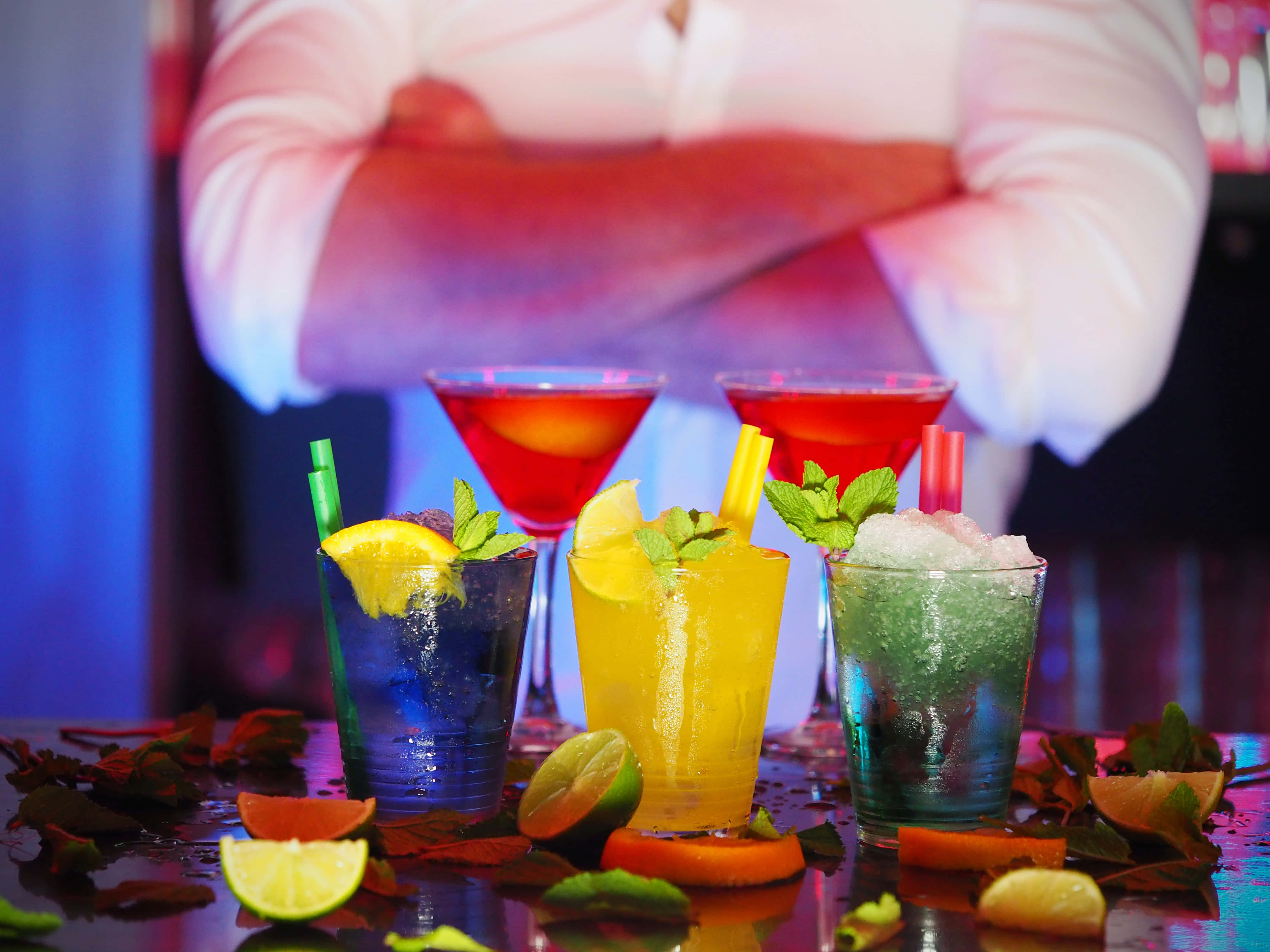 Summer Drinks 桌布 壁紙 — 色彩繽紛的雞尾酒放在彩色玻璃杯中，飾以柑橘和薄荷，置於霓虹燈下的吧檯上，背景有調酒師。