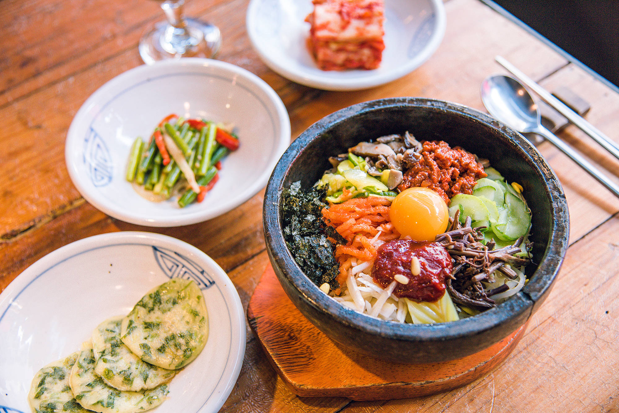 色彩繽紛的韓式拌飯與蔥餅套餐 壁紙 — 一個冒煙的石鍋韓國拌飯，上面放有生雞蛋和辣醬，旁邊有配菜，放在木桌上。