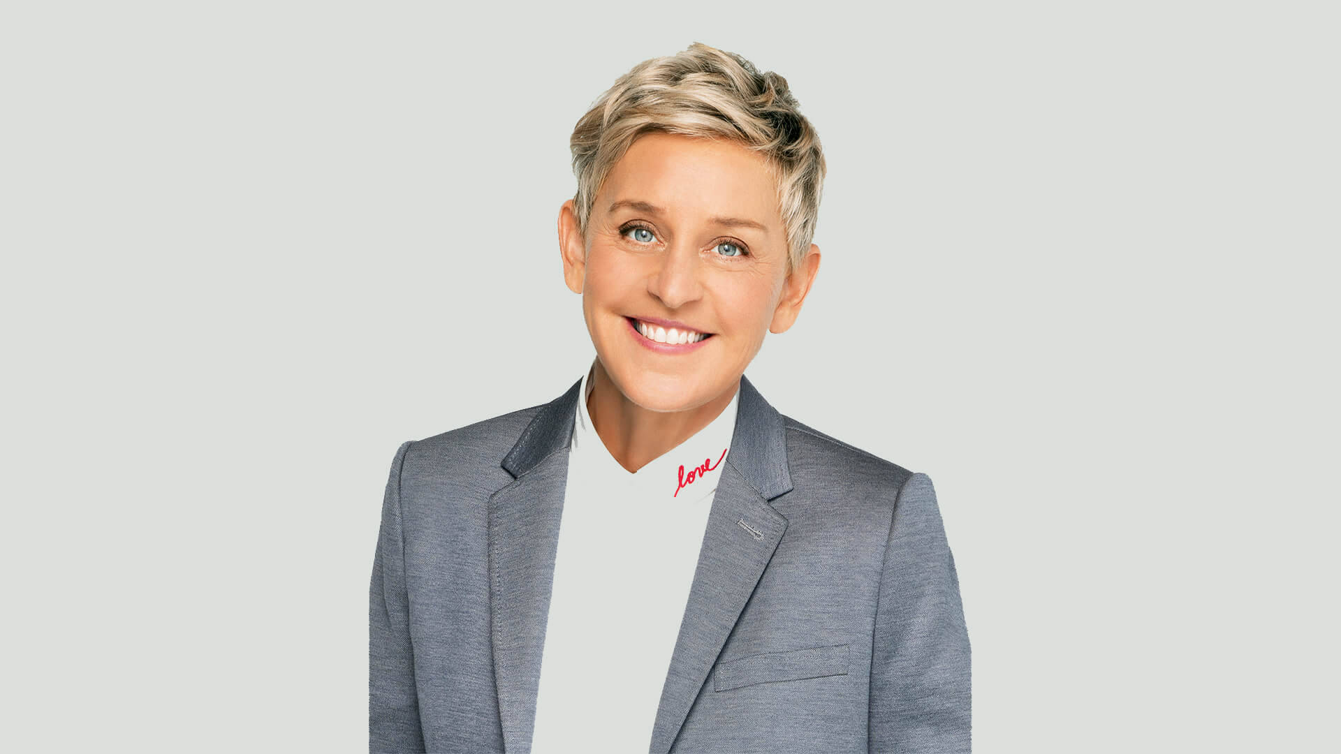 艾倫·德傑尼絲淺灰色西裝 壁紙 — Ellen DeGeneres 穿著灰色西裝外套配白色高領毛衣，紅色草書文字，溫暖地微笑。