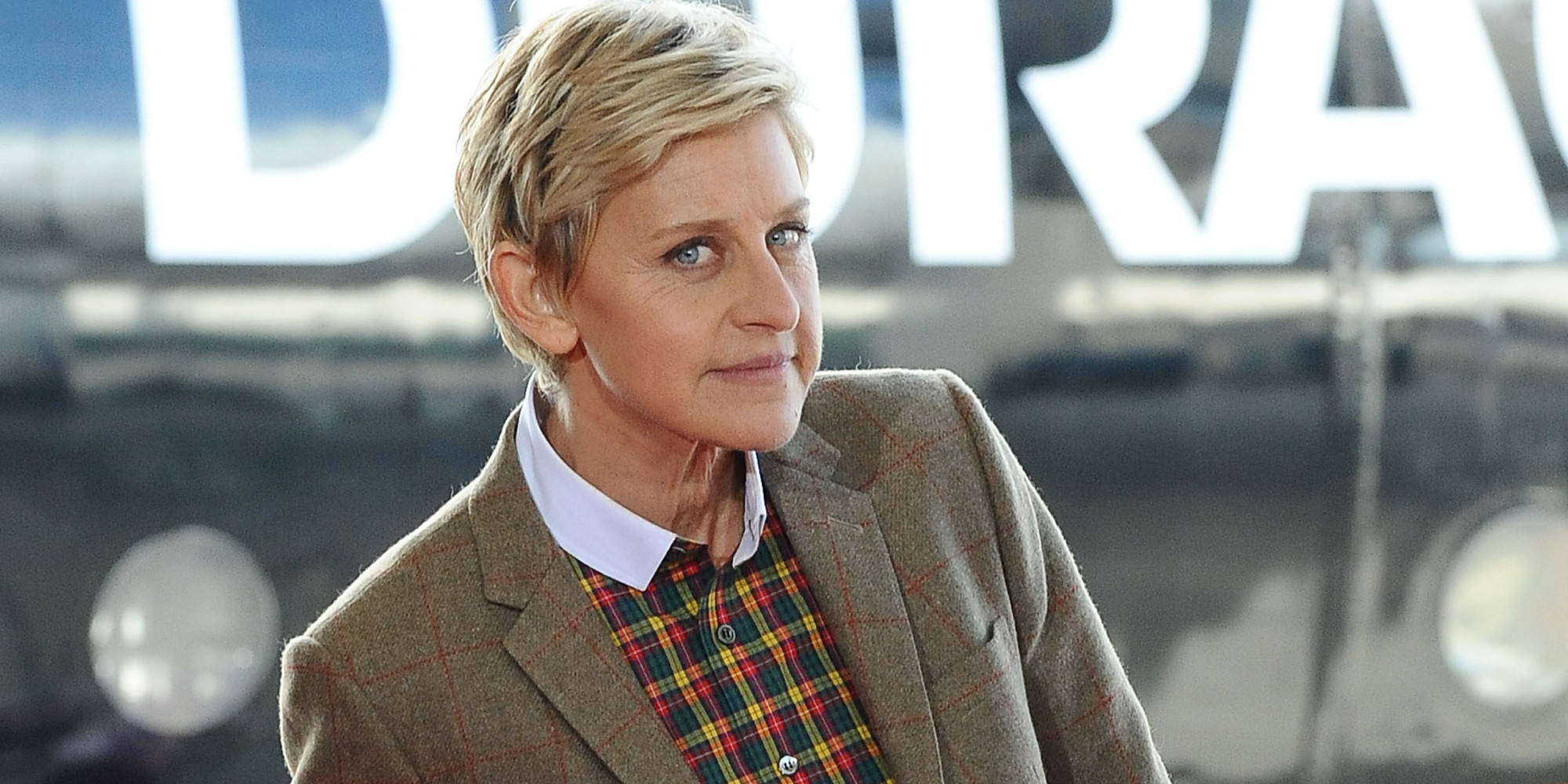 艾倫·德傑尼絲隨拍照片 壁紙 — Ellen DeGeneres 身穿棕色粗花呢外套和格仔恤衫，站在一輛帶有「DURACELL」標誌的模糊復古汽車前。