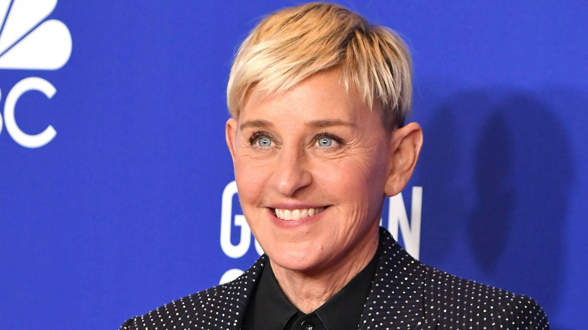 艾倫·德傑尼絲黑色波點西裝 壁紙 — Ellen DeGeneres 在活動中穿著黑色圓點西裝外套，在藍色NBC背景下溫暖地微笑。