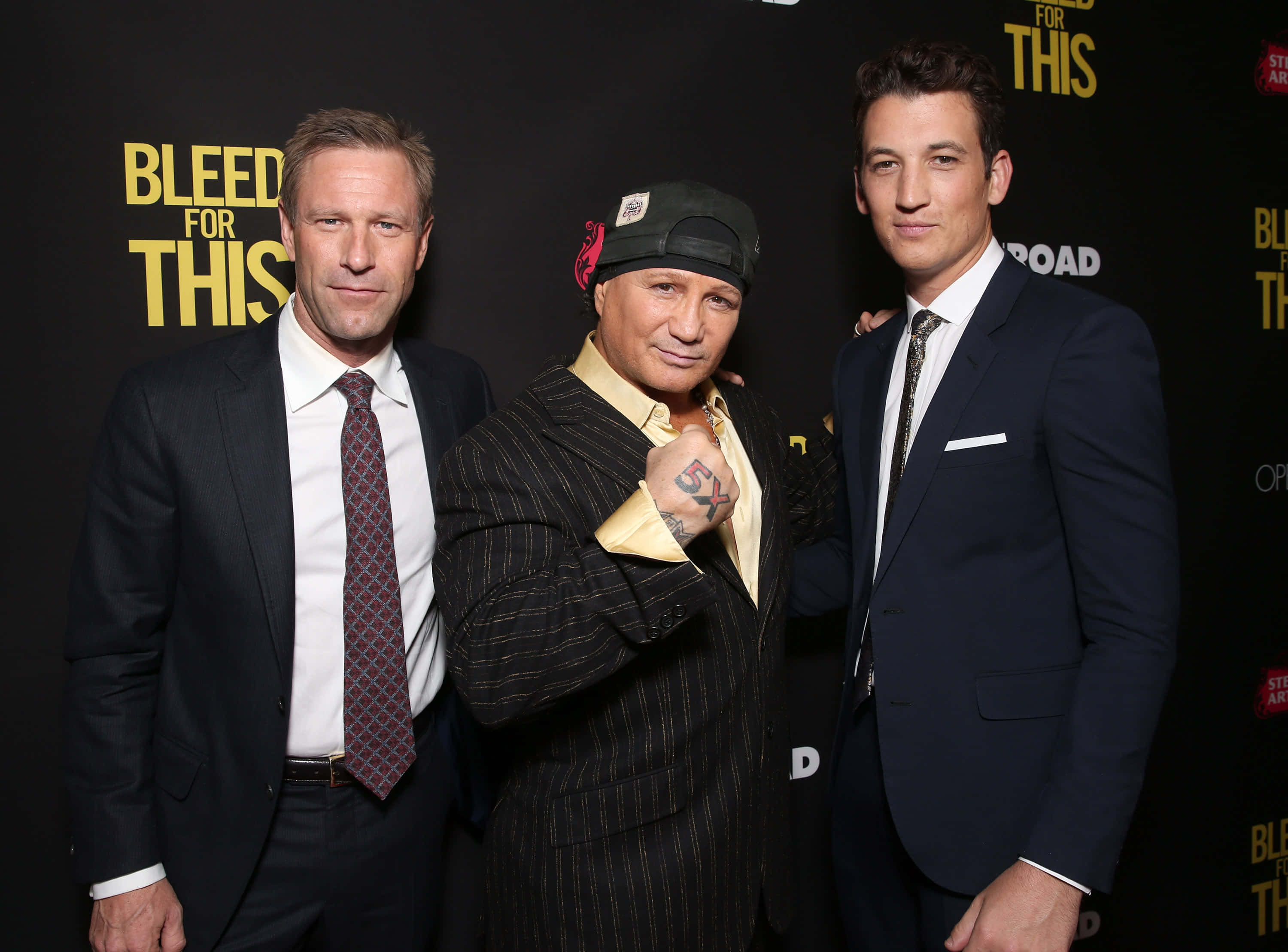 艾倫·艾克哈特《為愛搏命》首映 壁紙 — 三位身穿西裝的男子在《Bleed for This》電影首映禮上自信地擺姿勢，背景為大膽的黃色文字。