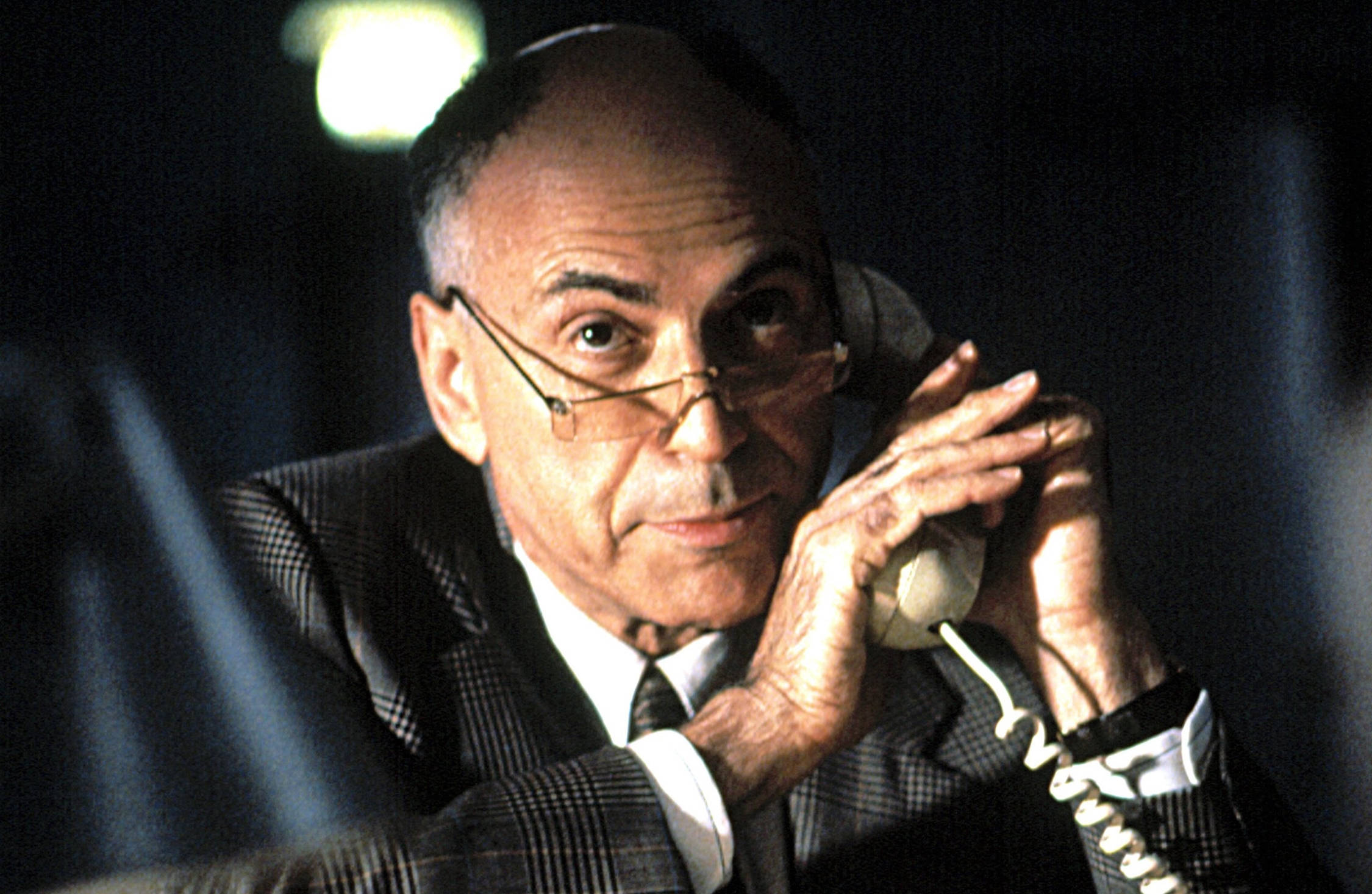 艾倫·阿金在電影《Glengarry Glen Ross》中飾演喬治·亞倫諾 壁紙 — 一個光頭戴眼鏡的男人在昏暗的房間裡，頭頂只有一盞燈，拿著一部復古電話貼在耳邊。