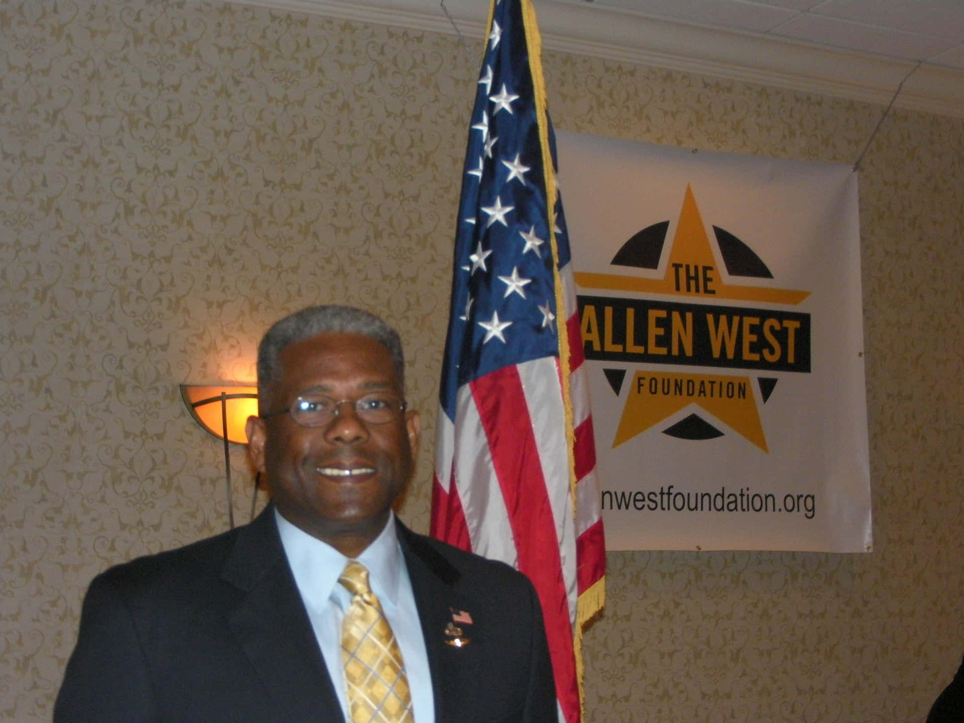活動中穿著西裝嘅男士微笑，背後有美國國旗同Allen West Foundation橫額。