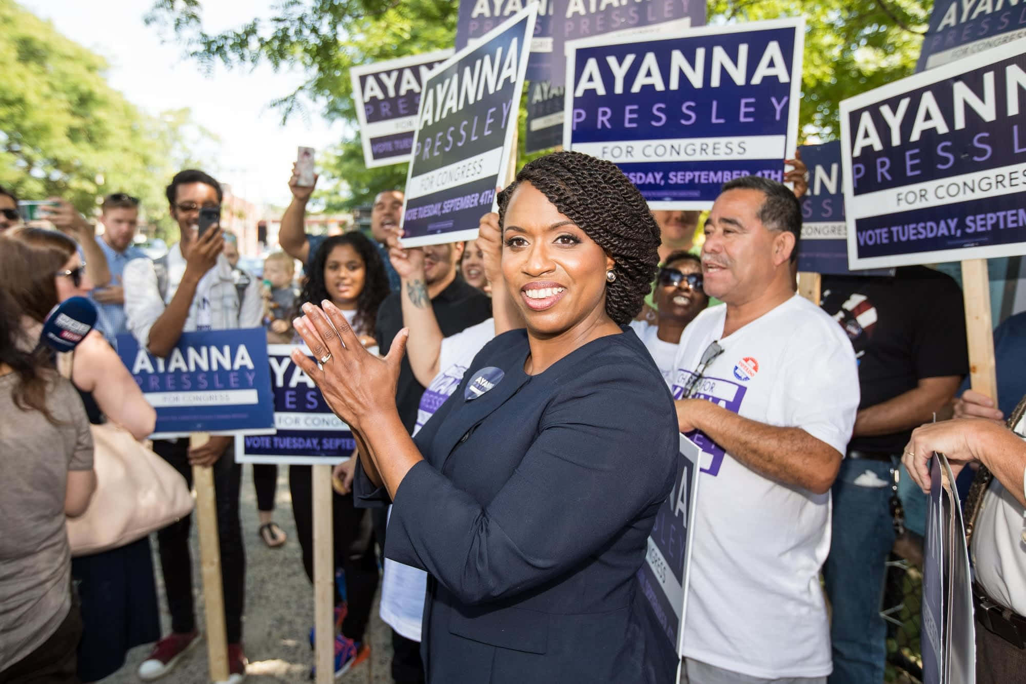 艾安娜·普雷斯利在競選活動中熱情鼓掌 壁紙 — Ayanna Pressley 在陽光明媚的戶外競選集會上微笑鼓掌，周圍是舉著她標語牌的支持者。