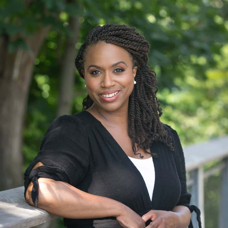 艾安娜·普雷斯利戶外自信擺姿 壁紙 — 一位紮辮子的微笑女士，在翠綠森林背景下隨意靠在石欄杆上。