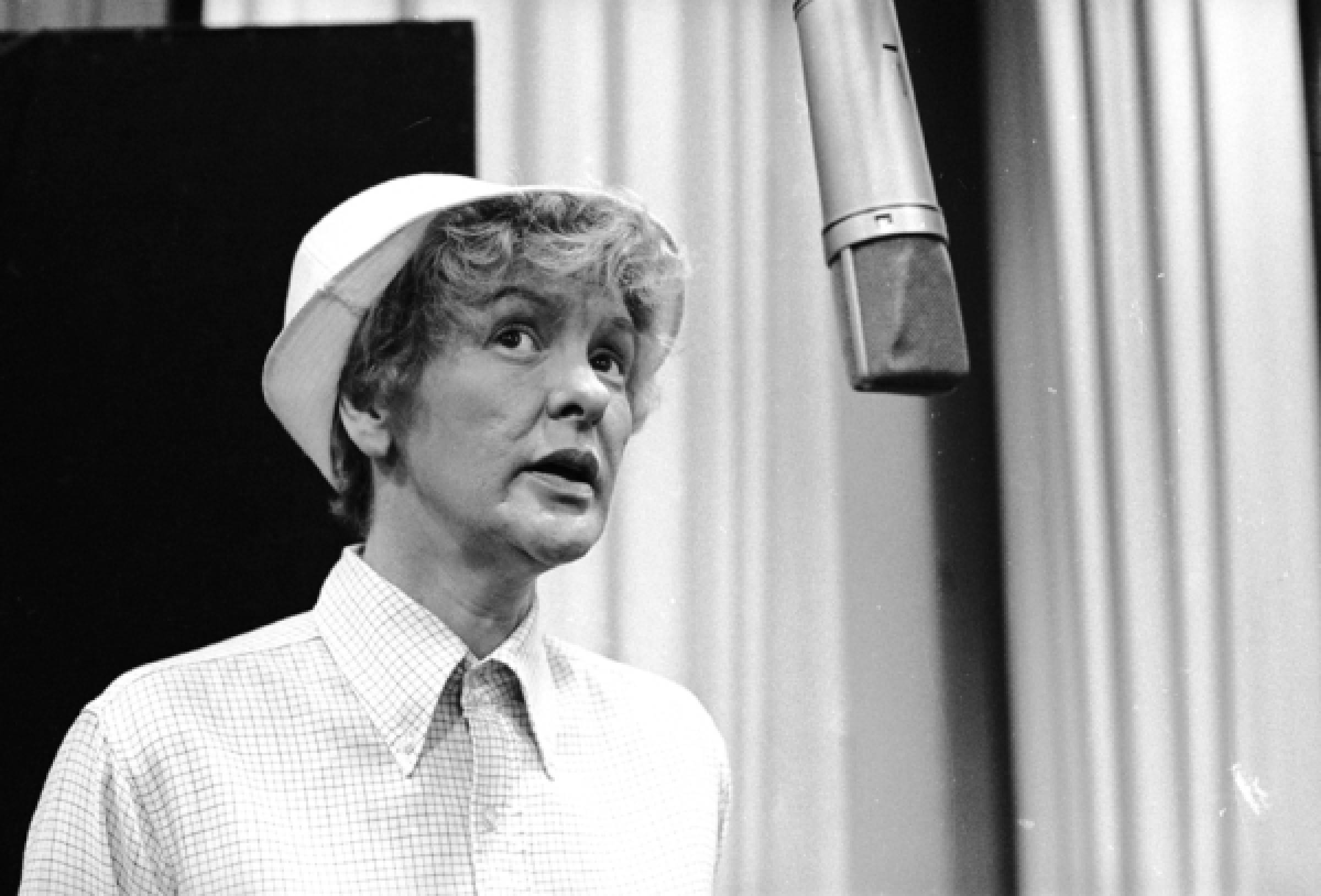 艾琳·斯特里奇在音樂劇《Forbidden Broadway》中演出 壁紙 — 一位戴白色帽子和格仔衫嘅女人喺有窗簾嘅錄音室入面向住復古咪高峰唱歌。