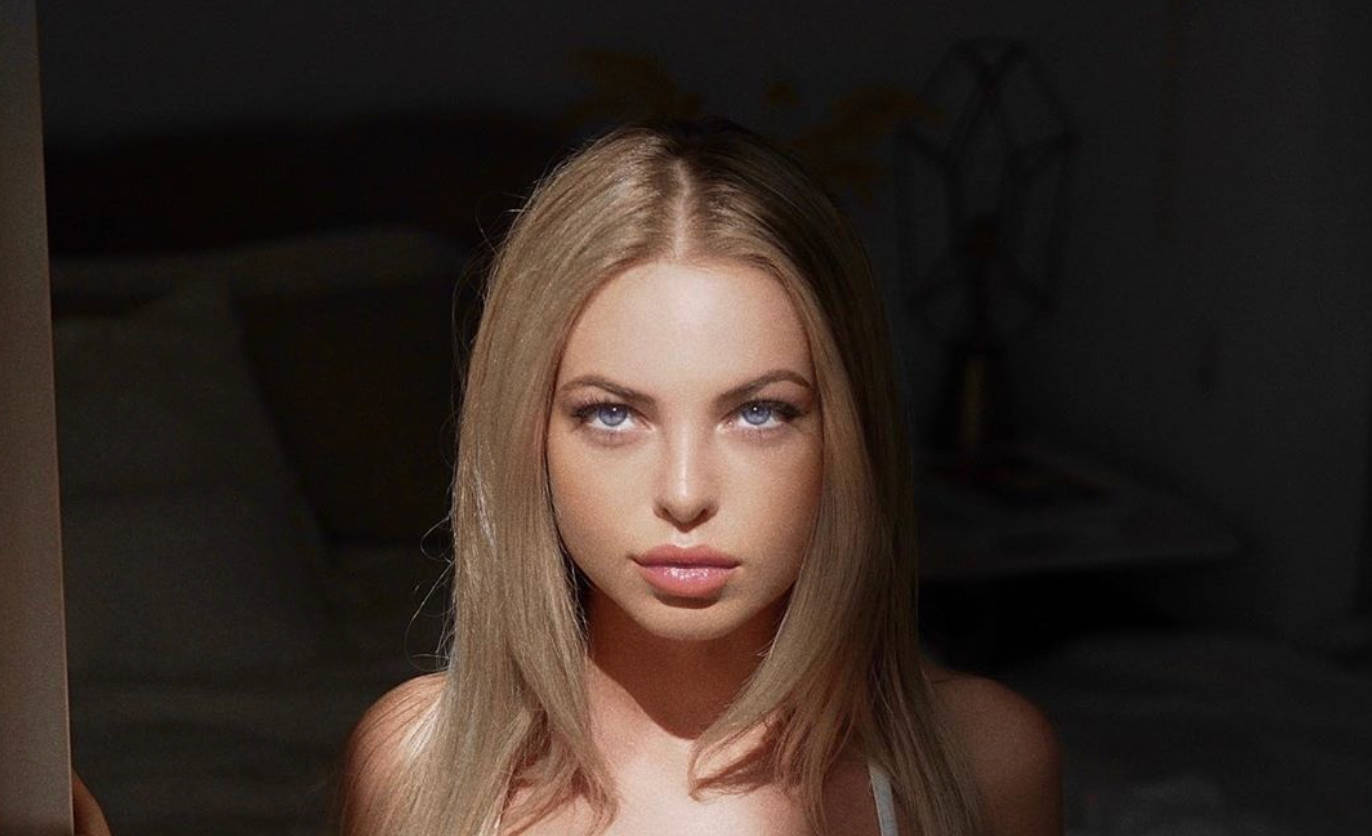 一位金髮碧眼的女性在光線昏暗的房間中直視鏡頭。