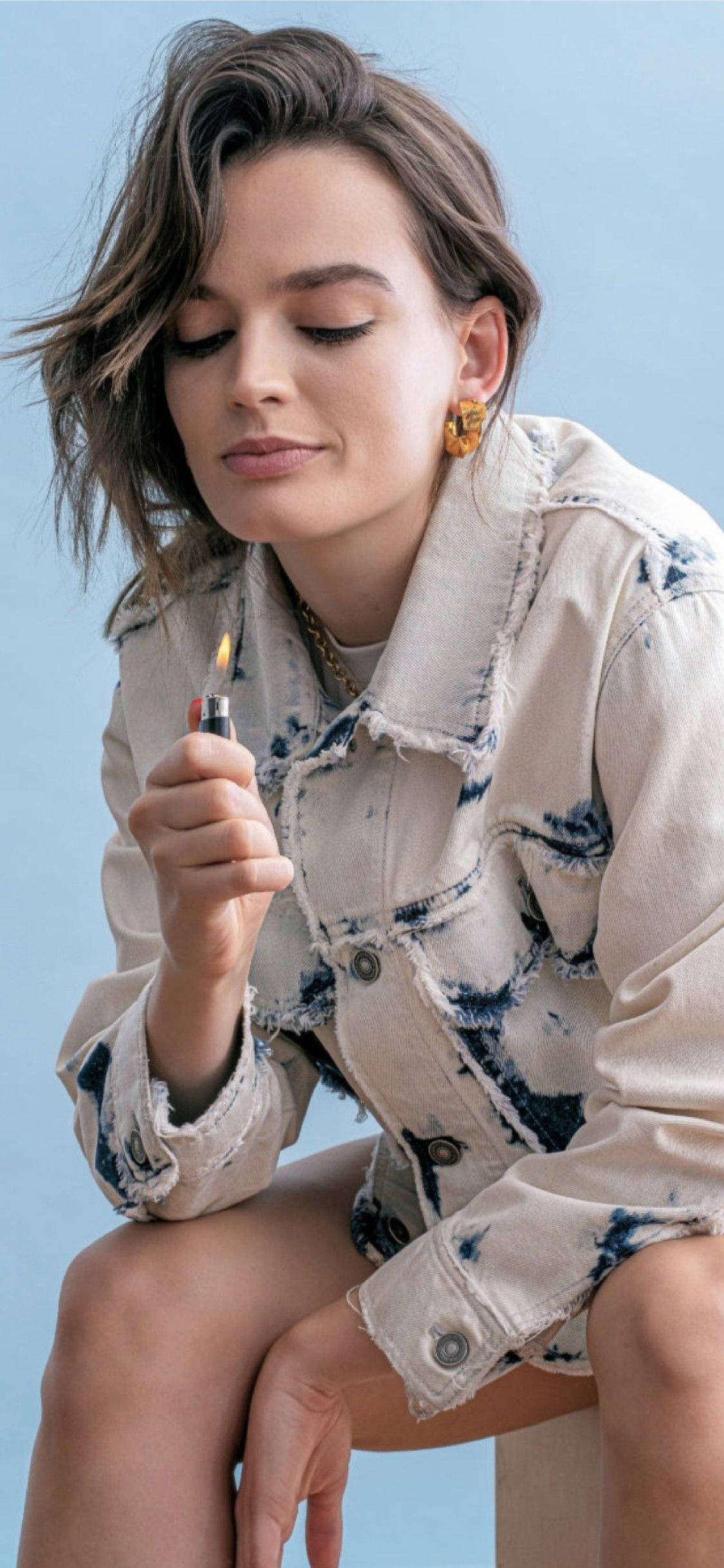 艾瑪·麥基手持打火機 壁紙 — 一位頭髮蓬亂的棕髮女性，平靜地坐在柔和的藍色背景前，手持點燃的打火機。