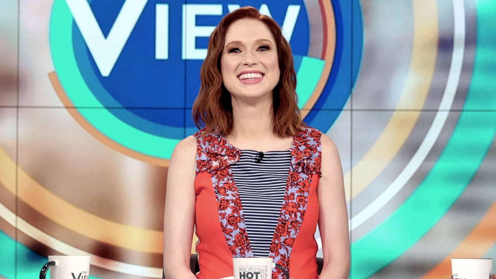 艾莉·凱默《The View》節目現場 壁紙 — 一位微笑的紅髮女性坐在脫口秀桌前，背景是色彩繽紛的圓形，帶有「VIEW」標誌。