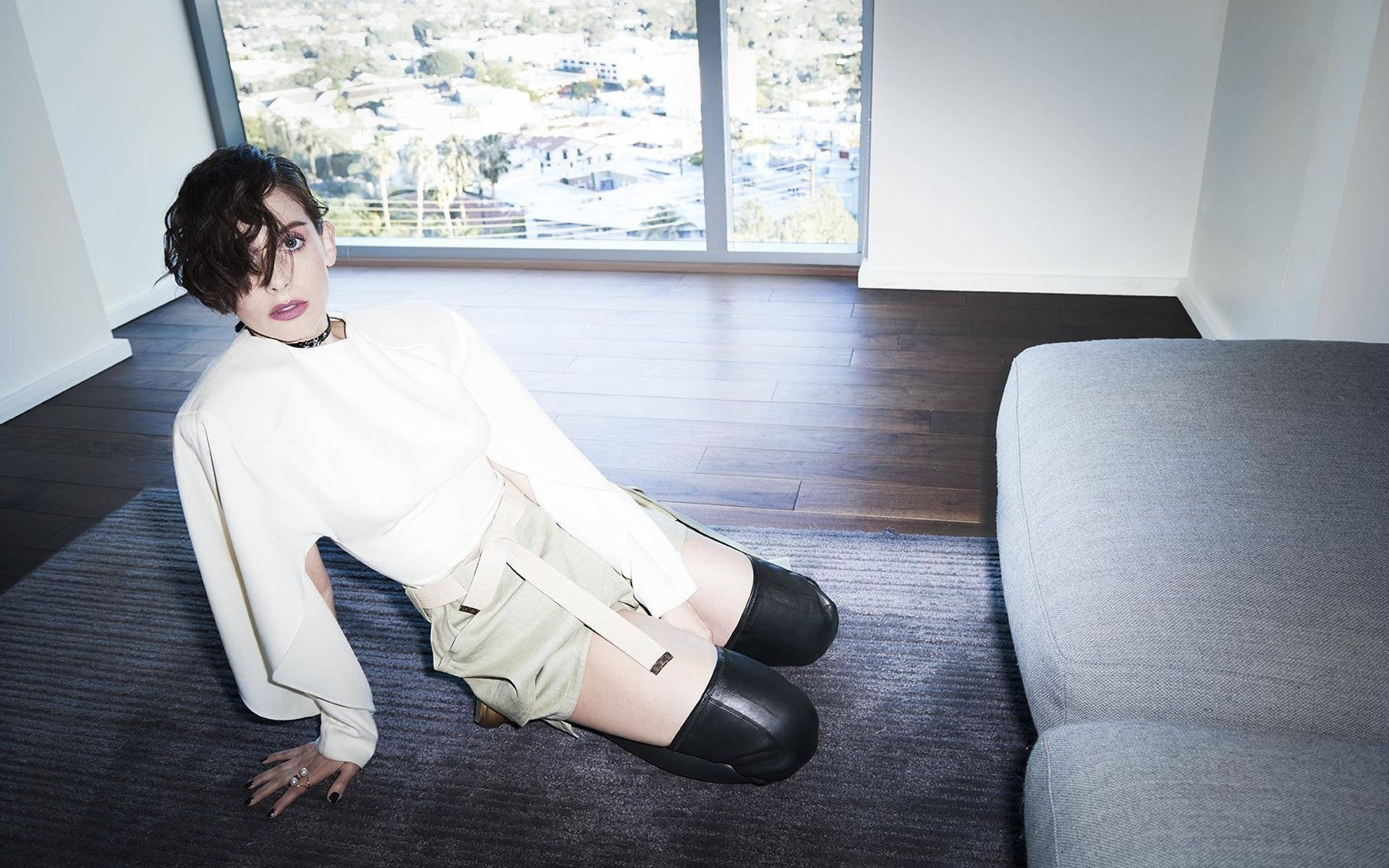 艾莉森·布里身穿白色上衣與黑色靴子跪地 壁紙 — 一位短黑髮女性坐在現代房間的條紋地毯上，穿著白色襯衫和黑色靴子，凝視鏡頭，背後的大窗戶可見城市景觀。