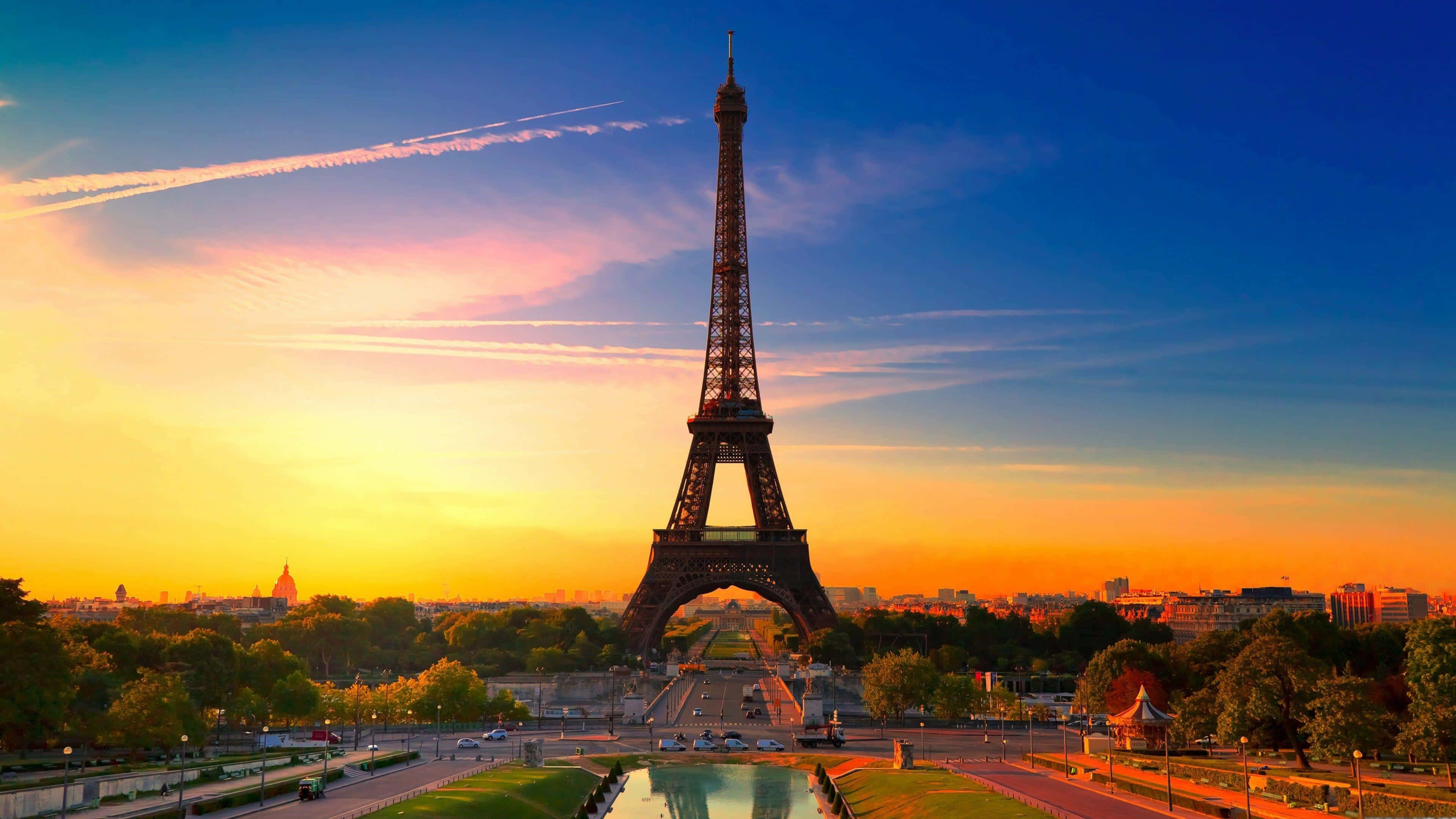 Eiffel Tower 桌布 壁紙 — 艾菲爾鐵塔在充滿活力的日落天空下高高聳立，金色的光芒照亮了巴黎的天際線和周圍的綠地。
