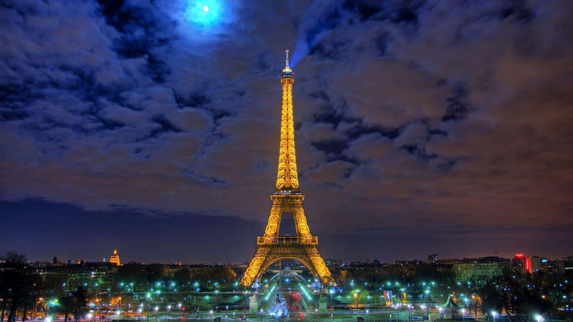 Eiffel Tower 桌布 壁紙 — 艾菲爾鐵塔在戲劇性的夜空下閃耀金色，伴隨著明亮的藍色月亮和散落的雲朵。