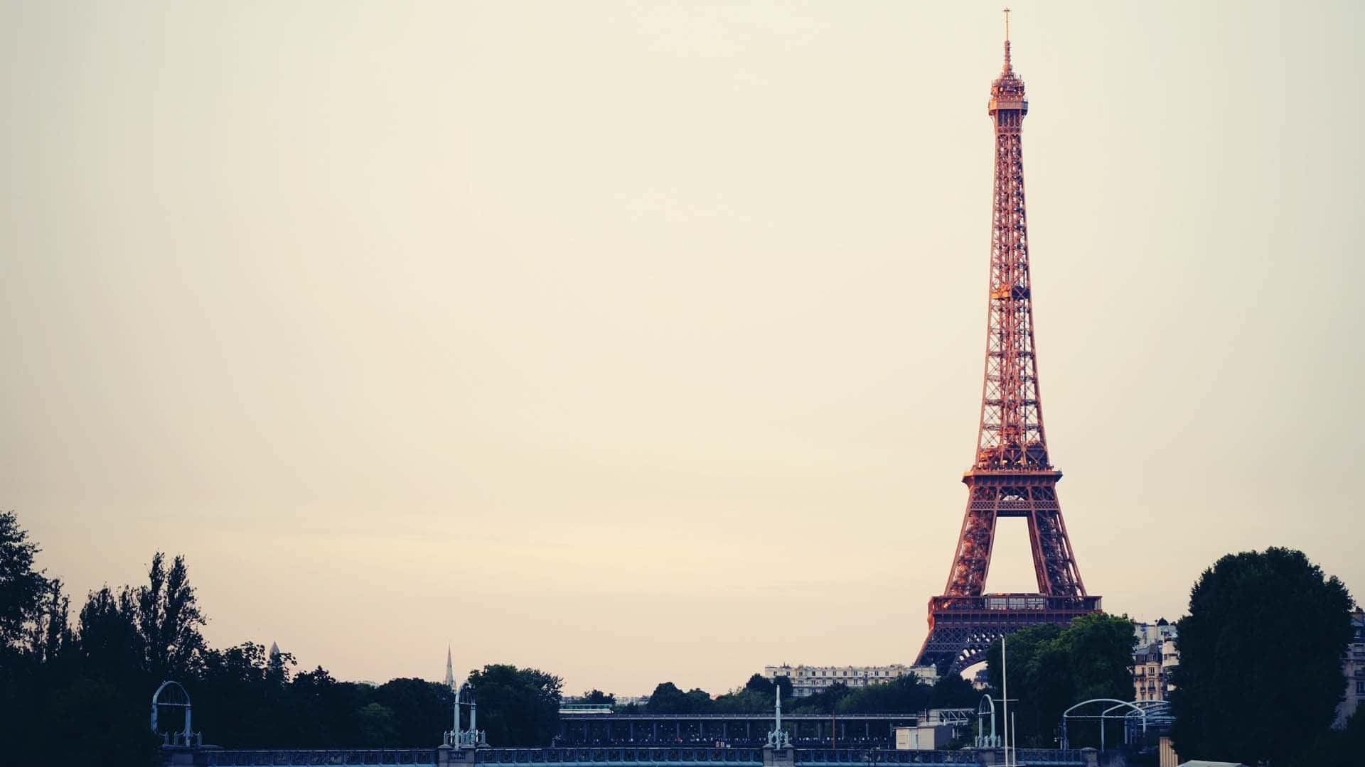 french 桌布 壁紙 — 艾菲爾鐵塔在柔和的粉彩天空下高聳，暮色籠罩巴黎，樹木和遠處建築物形成剪影。