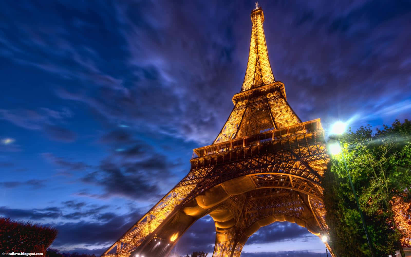 Eiffel Tower 桌布 壁紙 — 艾菲爾鐵塔在深藍色雲朵和金色燈光照亮其複雜鐵結構的戲劇性暮色天空下溫暖地發光。