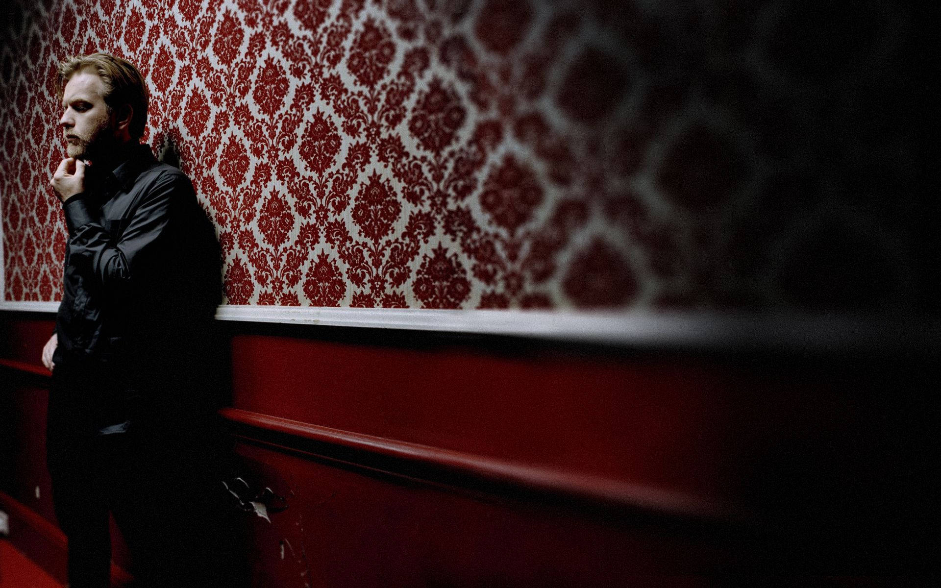 男人靠在一個光線昏暗的房間裡，牆上貼著紅白圖案牆紙。