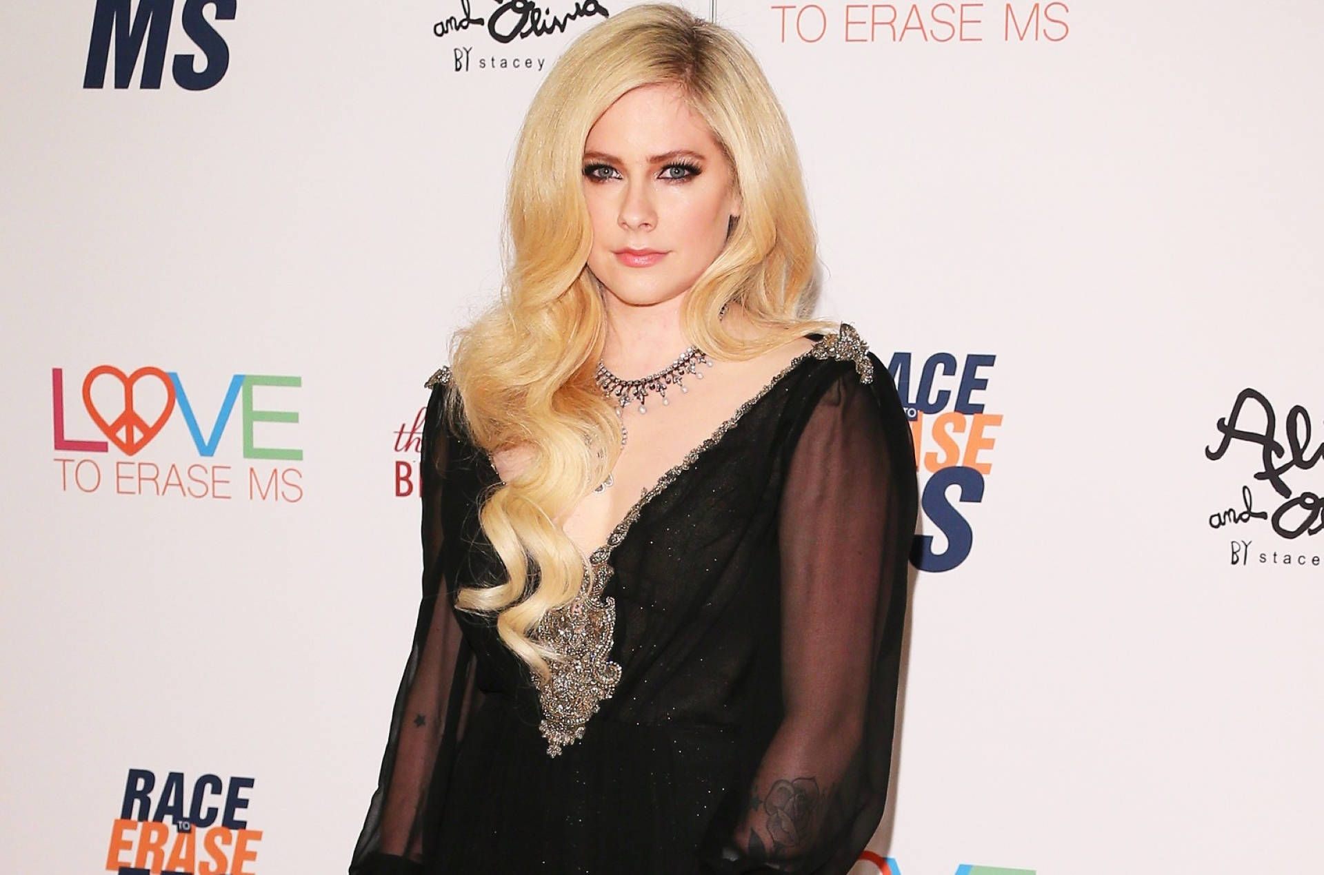 Avril Lavigne 在慈善活動中穿著黑色長袍，背景有品牌標誌。