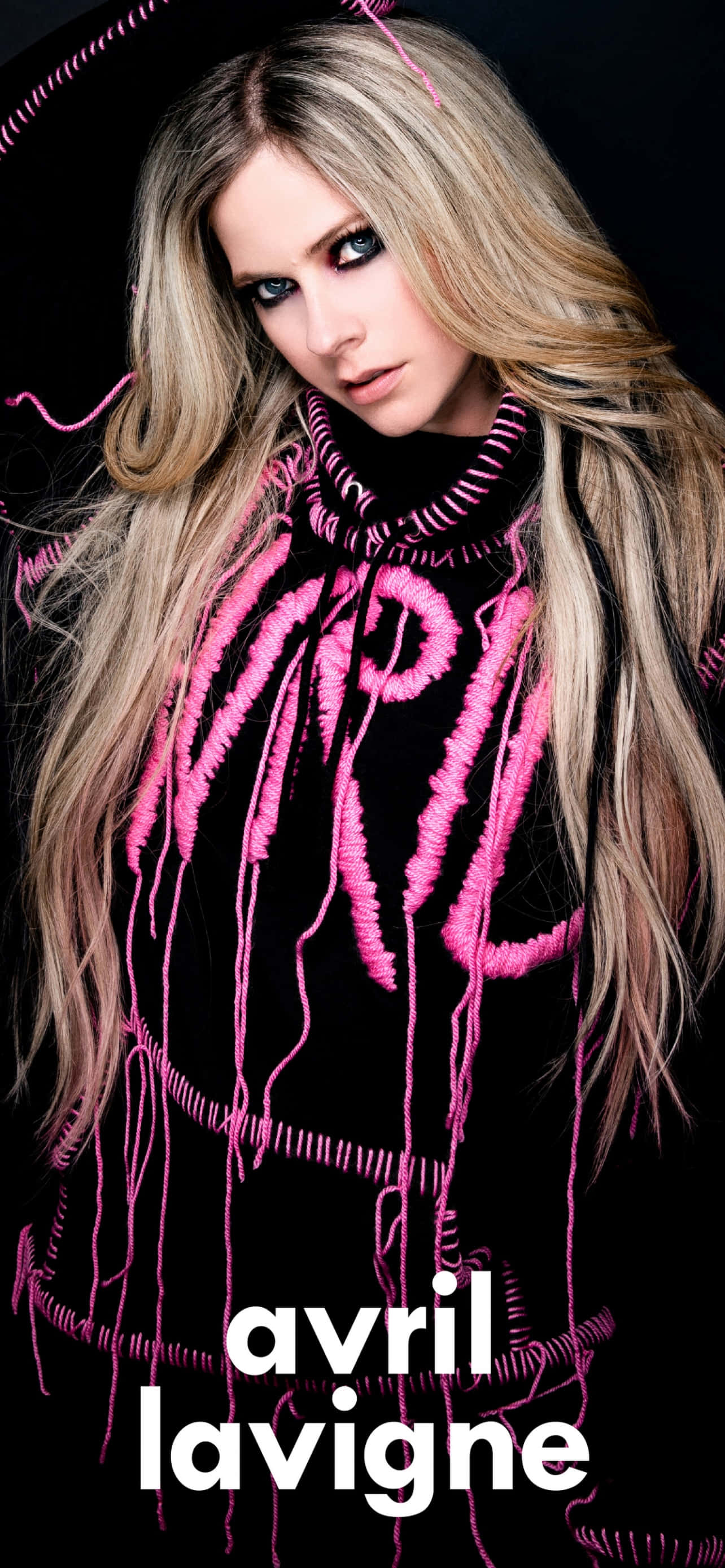 艾薇兒·拉維尼的時尚生活 壁紙 — Avril Lavigne 穿著黑色高領毛衣，帶有醒目的粉紅色縫線字母和流蘇，在黑暗背景下凝視。