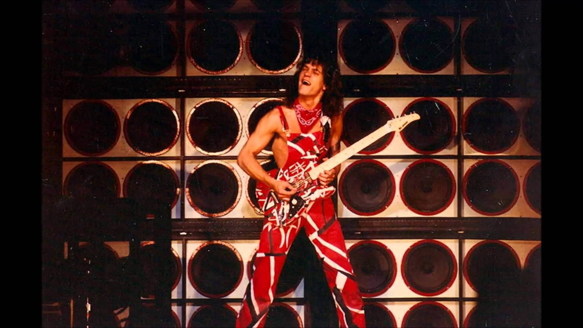 艾迪·范·海倫 Frankenstrat 連身衣 壁紙 — 一位身穿紅白條紋服裝的搖滾結他手，在圓形揚聲器牆前的舞台上彈奏相配的結他。